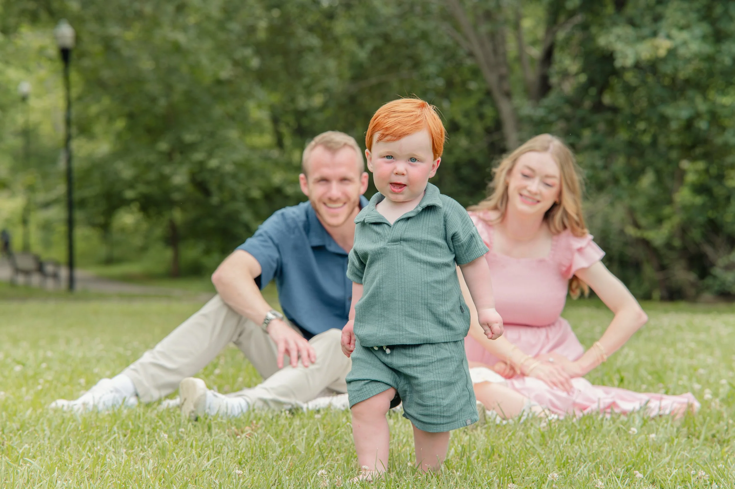 A young boy with red hair walking toward the camera on green grass, with a man and woman sitting behind him on a blanket in a park, surrounded by trees.