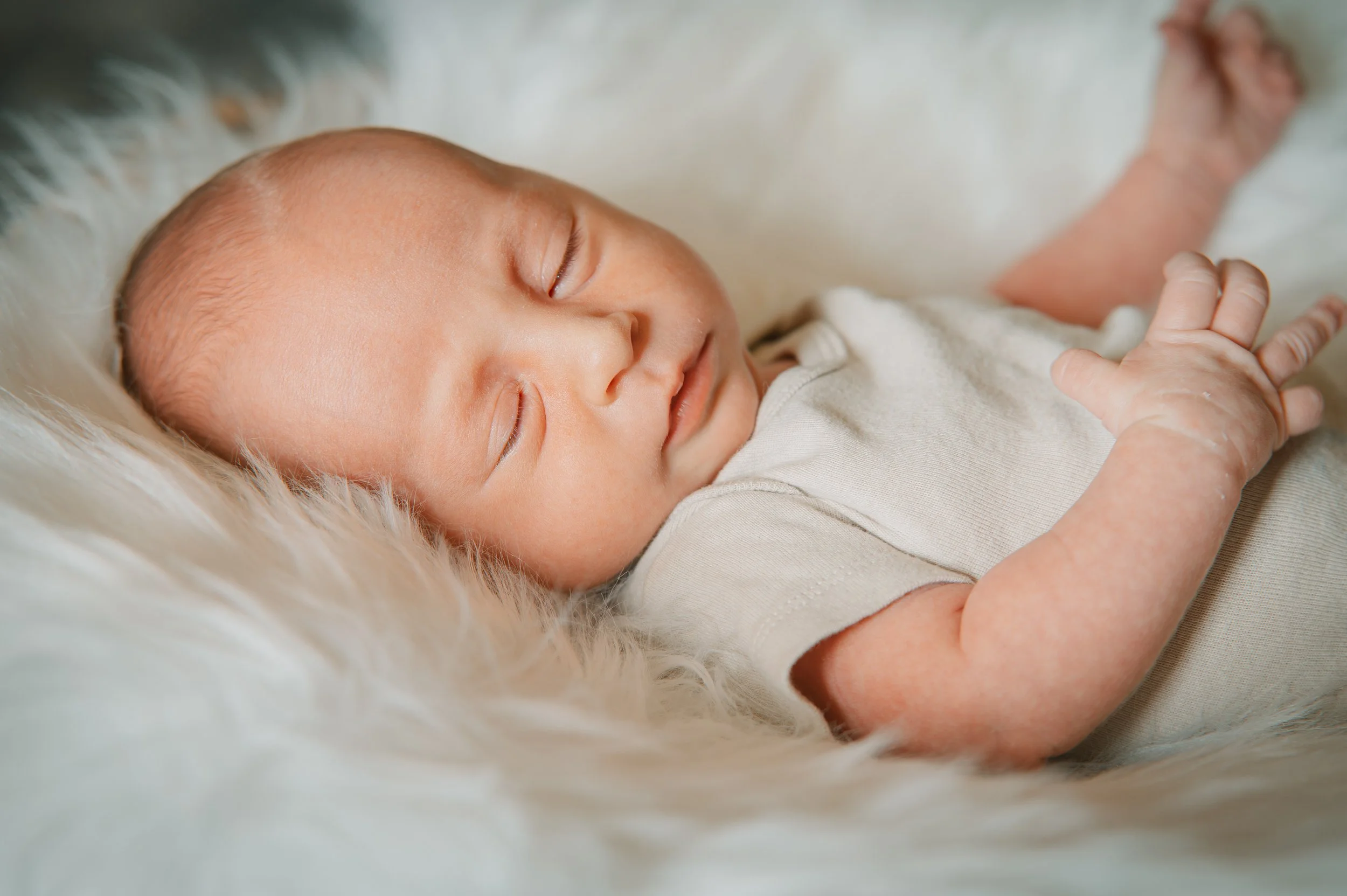Close-up of a sleeping newborn baby lying on a soft, furry white surface, wearing a light-colored onesie.