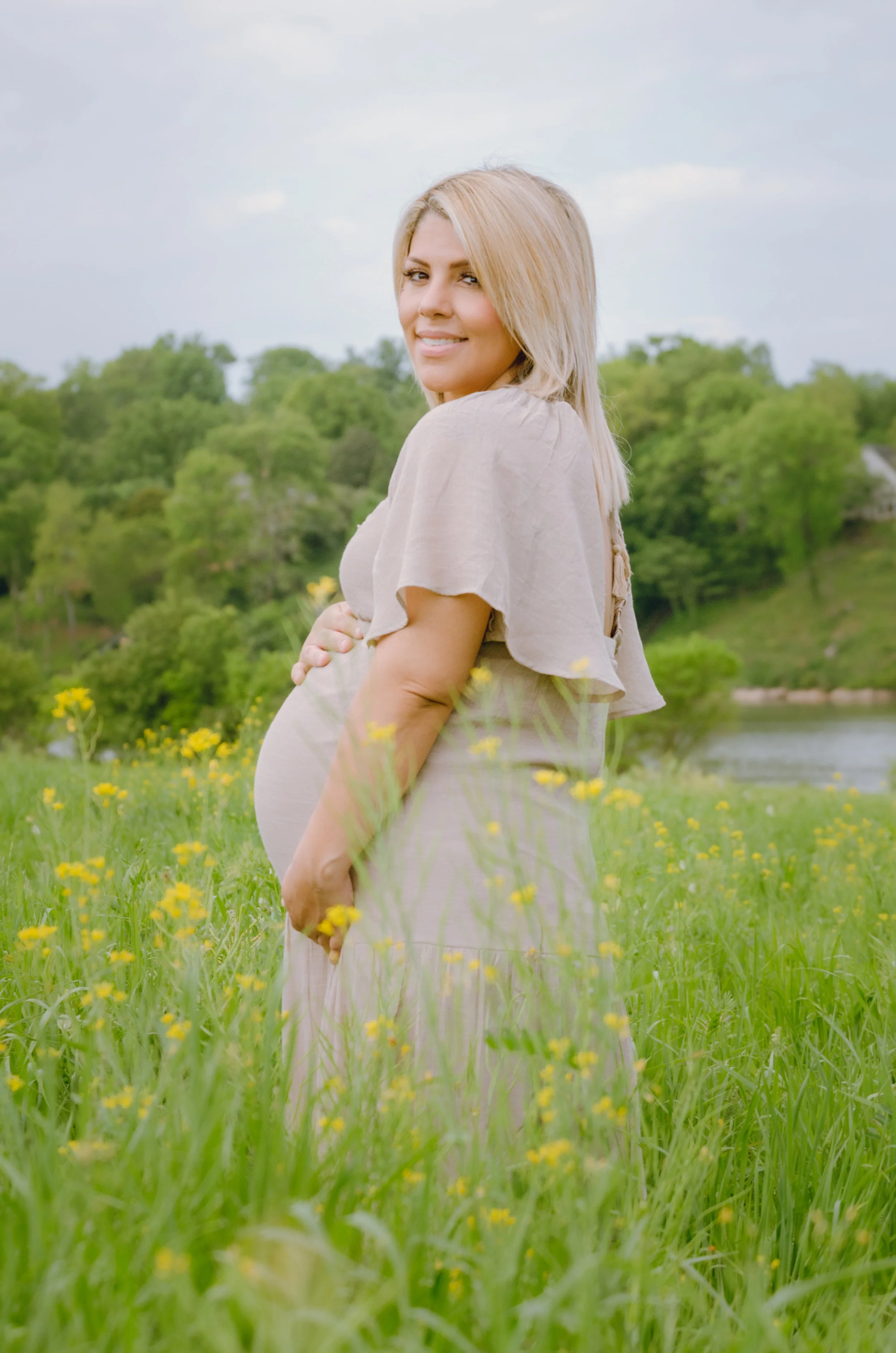 Pregnant woman with blonde hair standing in a grassy field with yellow flowers, green trees, river, and cloudy sky in the background, smiling and looking over her shoulder.