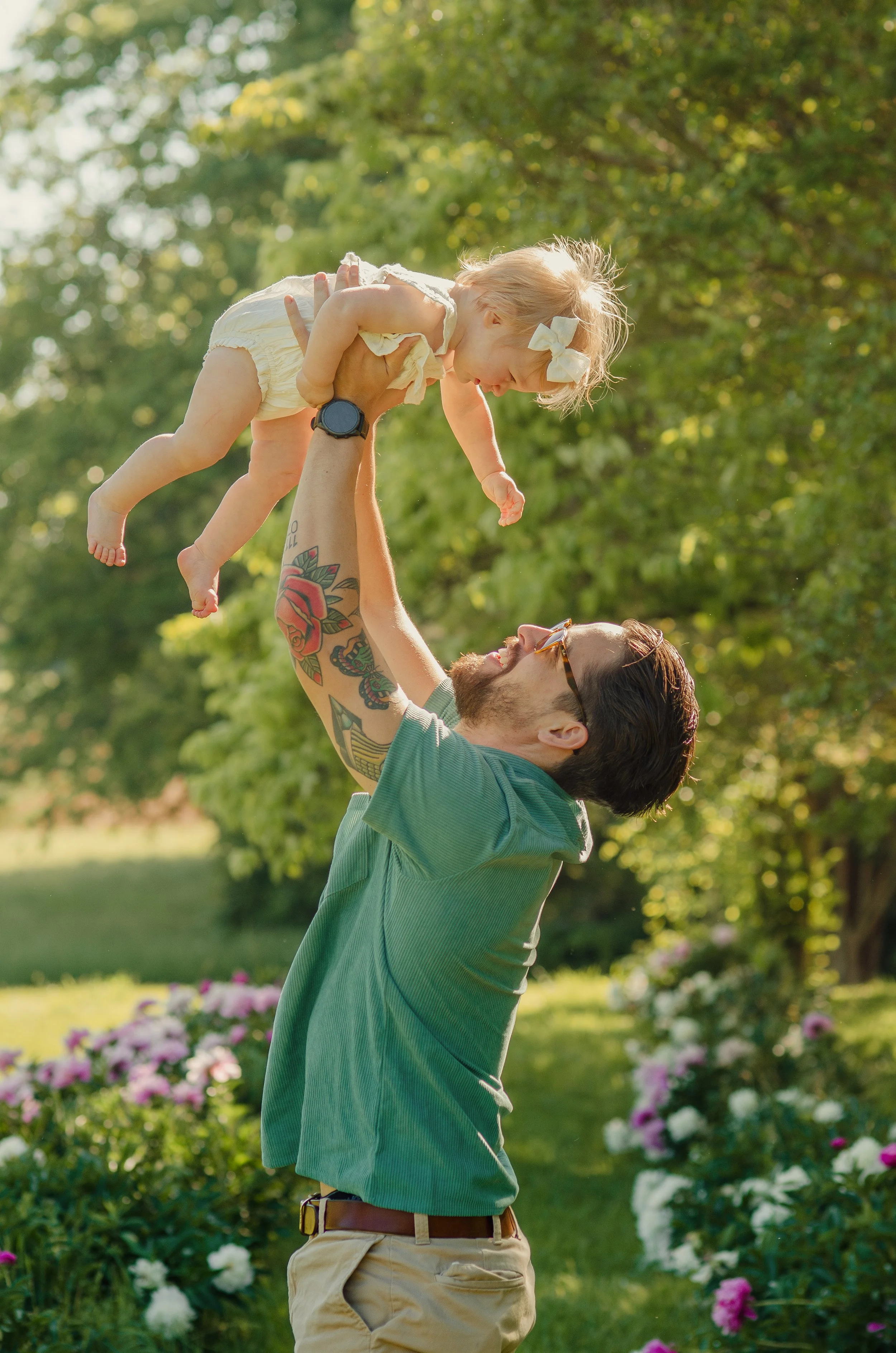 A man with tattoos wearing sunglasses and a green t-shirt lifting a young girl with blond hair and a white bow in a park with trees and pink flowers in the background.