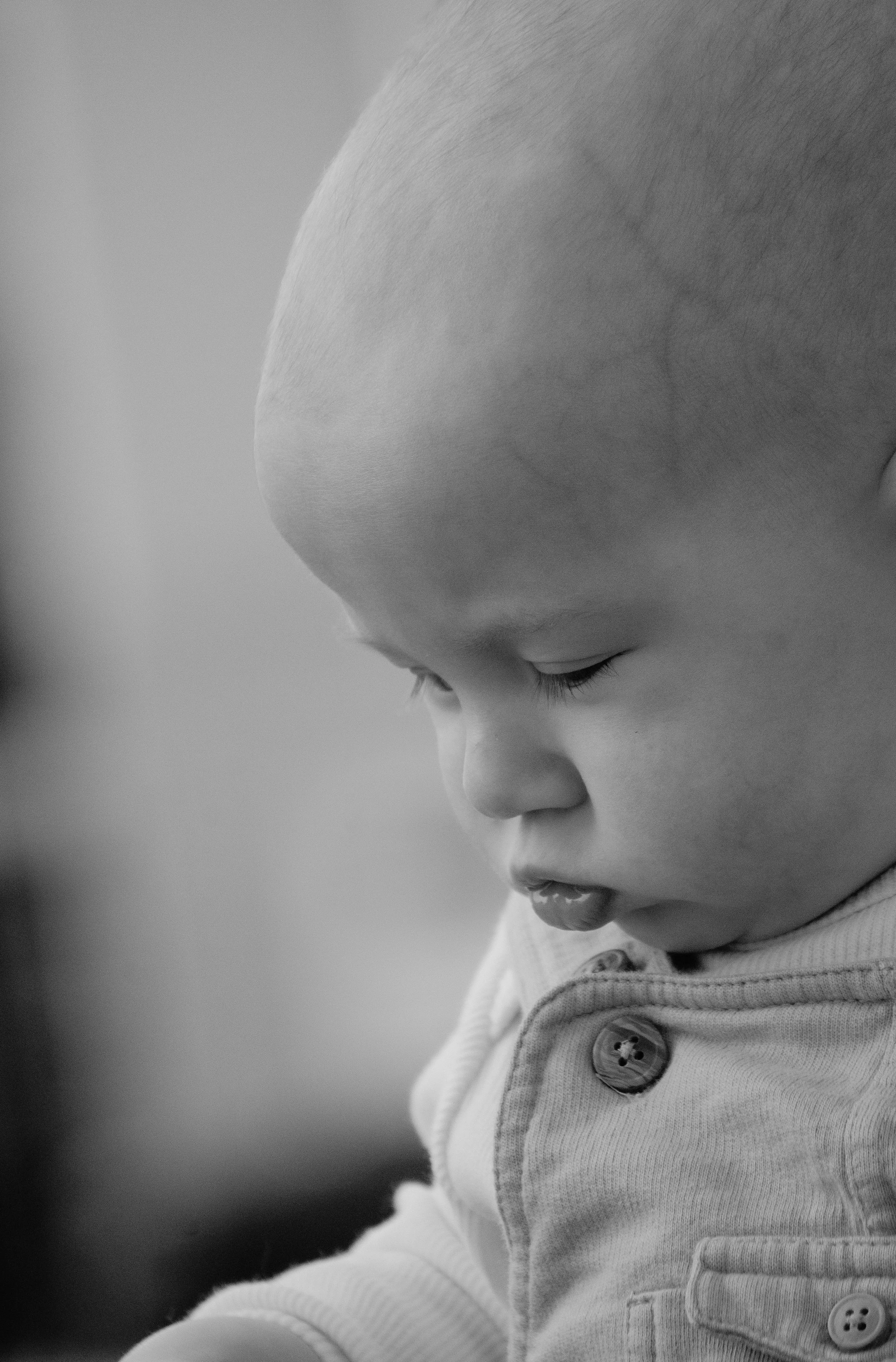 A young boy with a shaved head and a button-up shirt, looking downward with a calm expression.