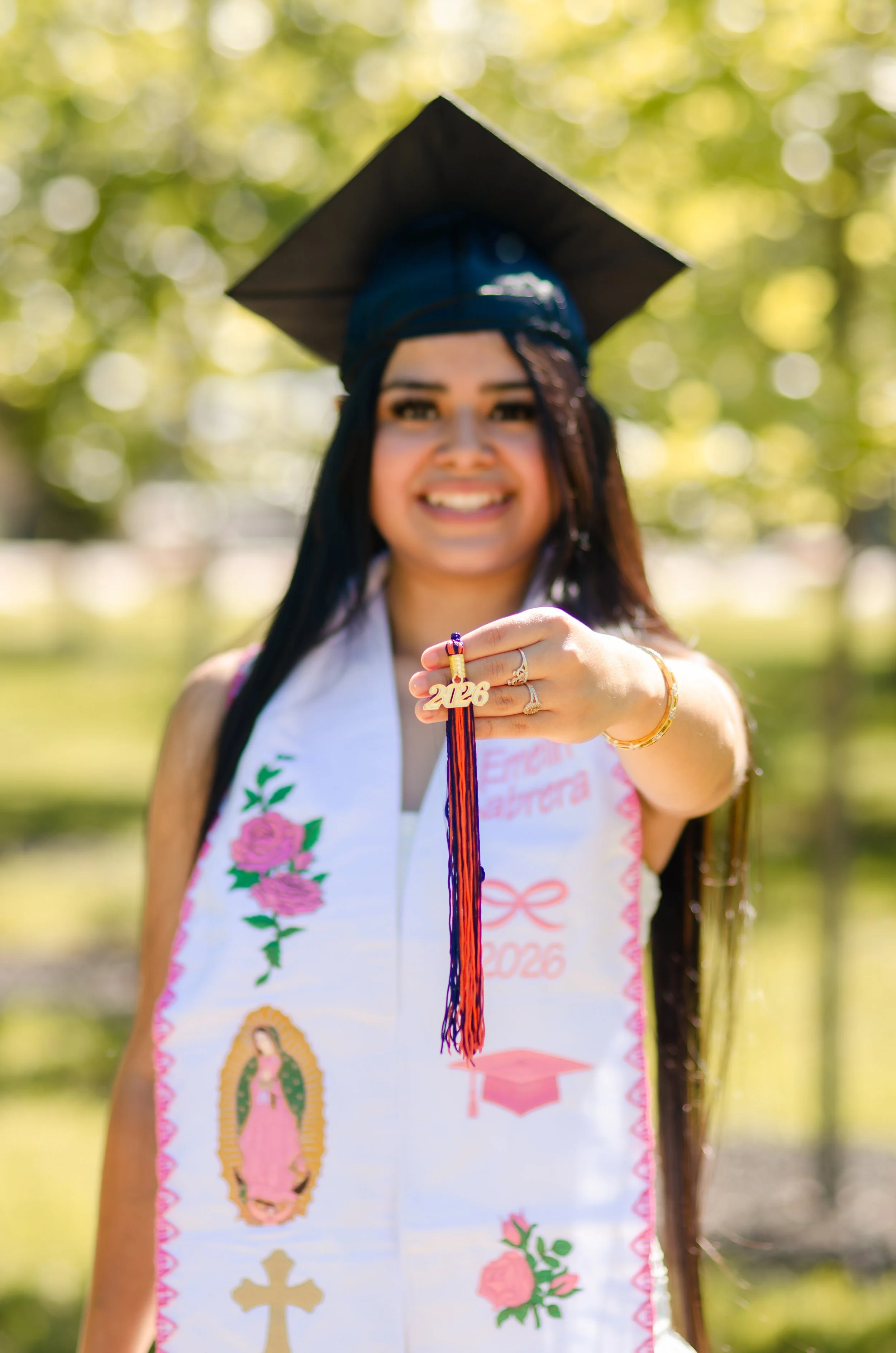 A young woman in a graduation cap and gown, smiling, outdoors with a green, blurred background, displaying her graduation medal and a sash with various symbols.