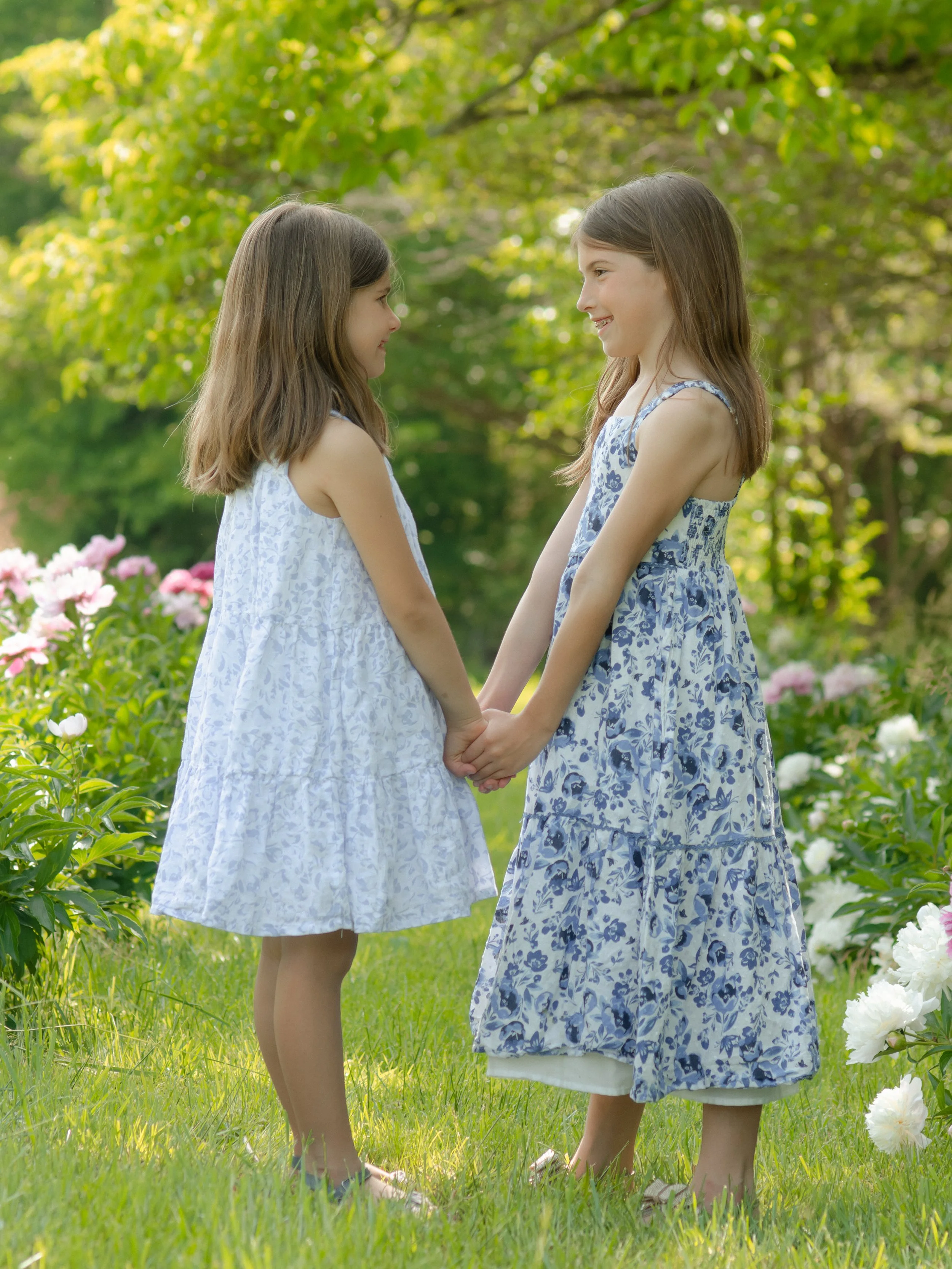 Knoxville photosession with two sisters holding hands and facing each other in a garden with trees and flowers, dressed in light blue and white floral dresses.