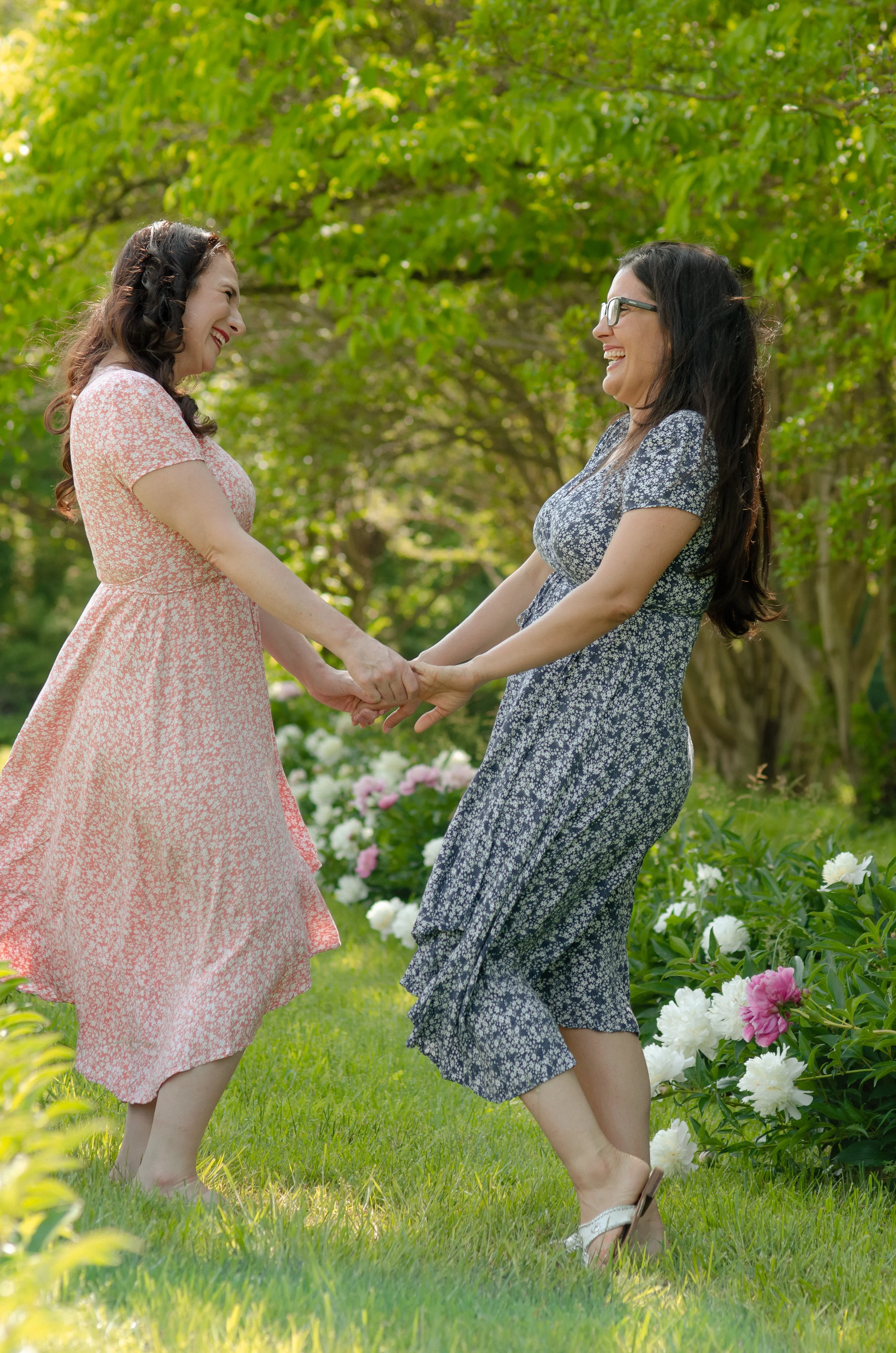 Photo session with adult sisters holding hands and smiling at each other outdoors in a garden with blooming peonies and green trees.