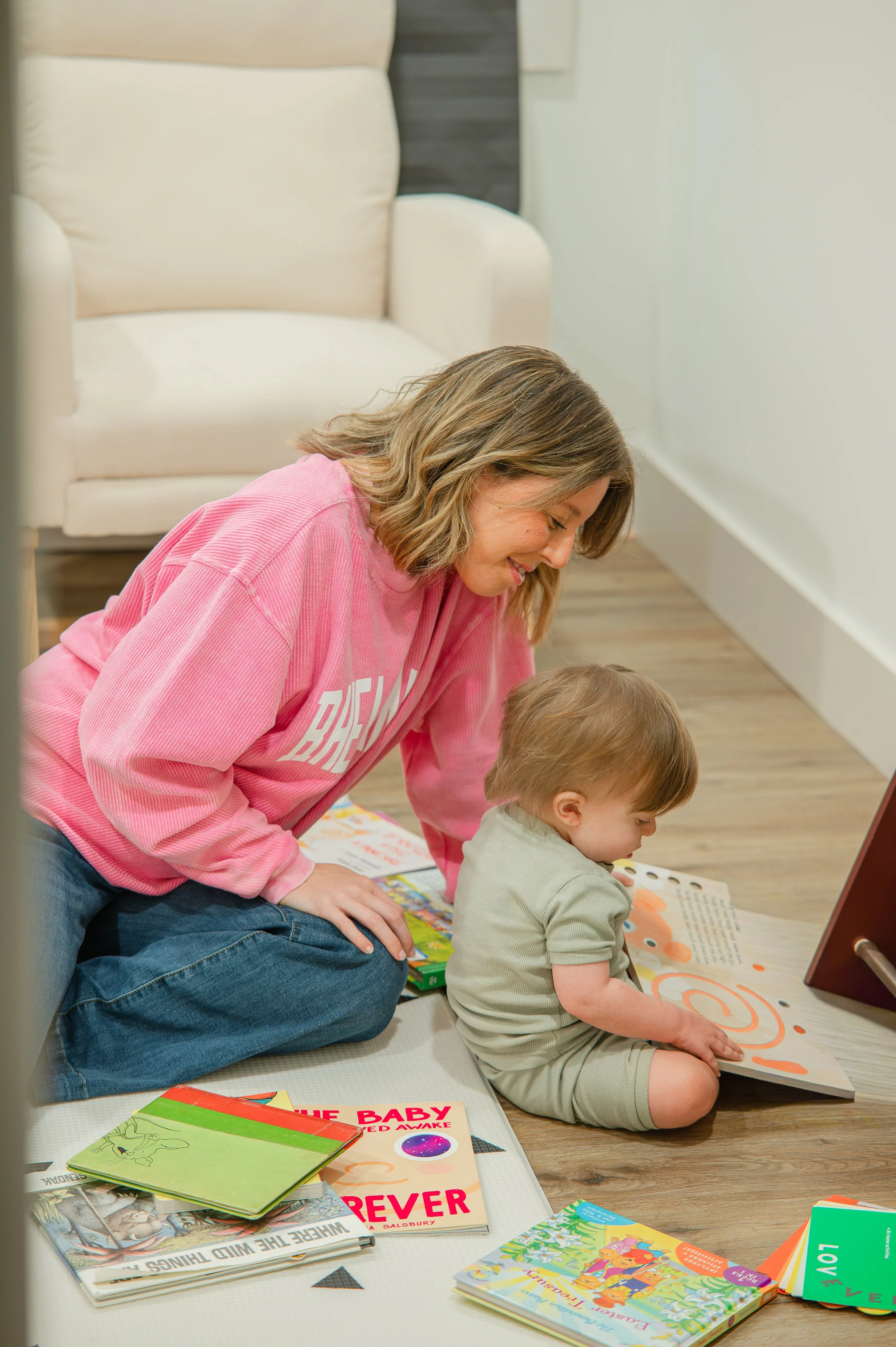 A woman and a young boy sitting on a floor surrounded by colorful children's books, engaging in reading or storytelling together. The woman is wearing a pink sweatshirt and the boy is wearing a light green shirt.