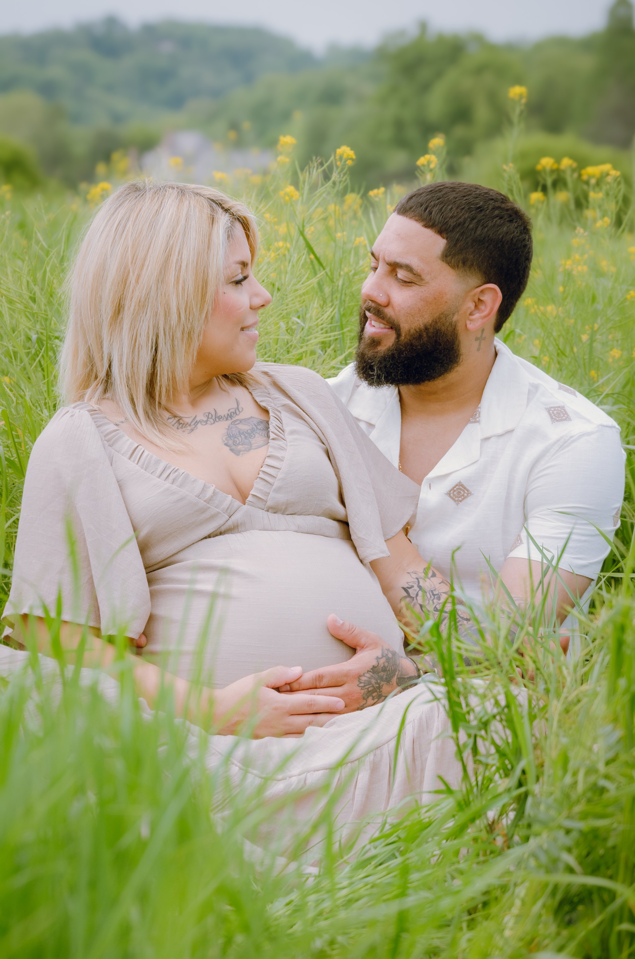 Knoxville maternity photo session, expecting couple sitting in a grassy field with yellow flowers, looking at each other lovingly.