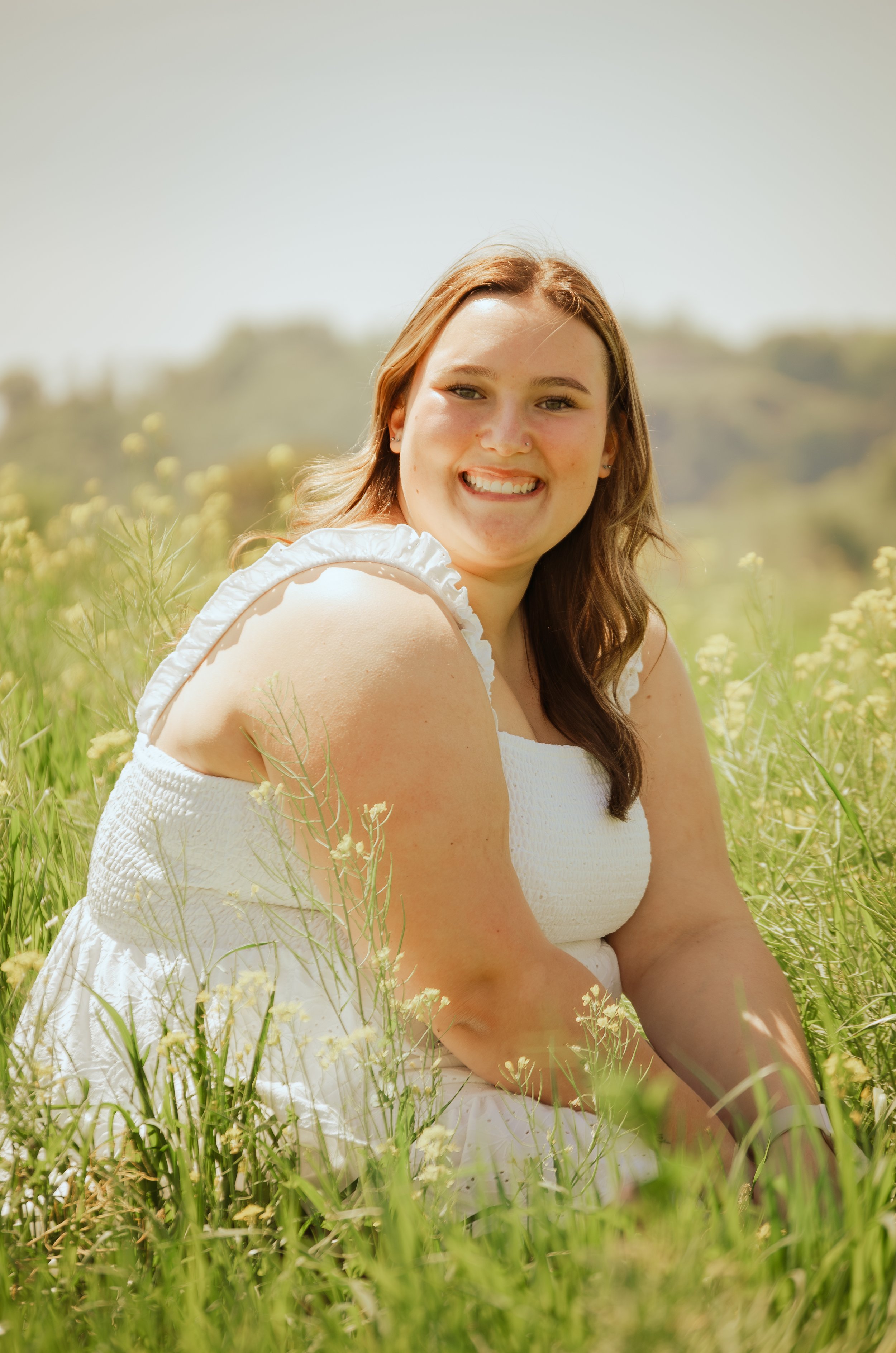 A young woman with long brown hair and a white dress sitting in a field of yellow flowers, smiling at the camera.