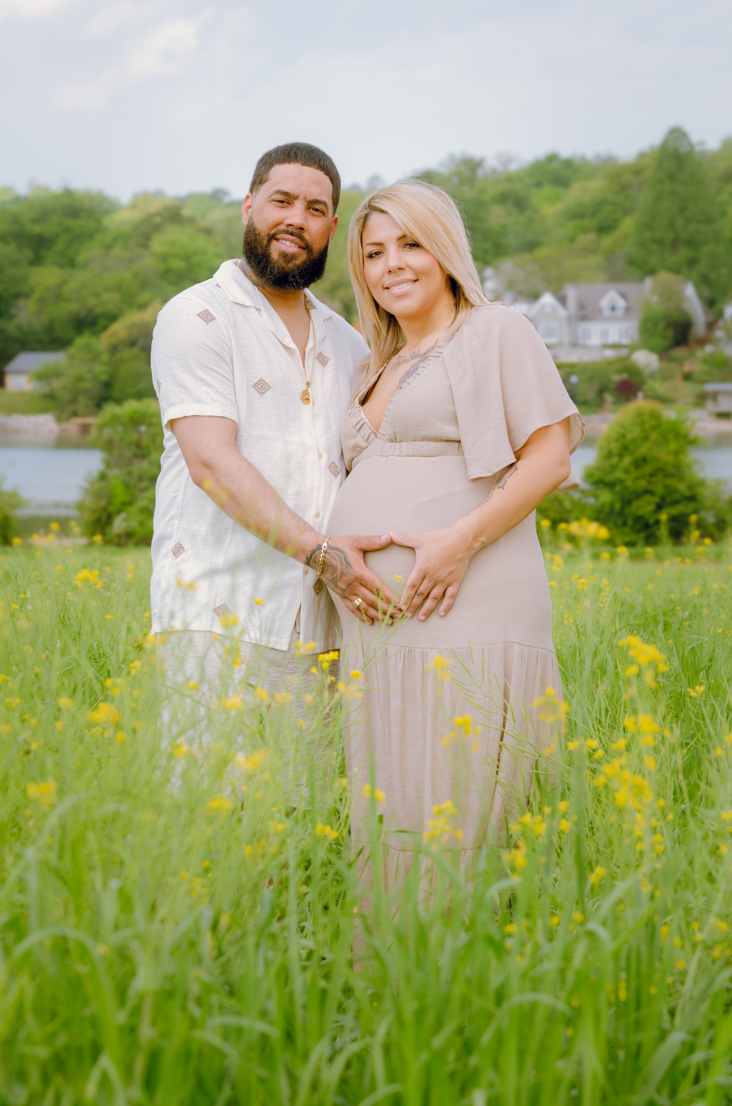 A pregnant woman and a man standing together in a grassy field with yellow flowers, holding hands on her belly, with trees and houses in the background.