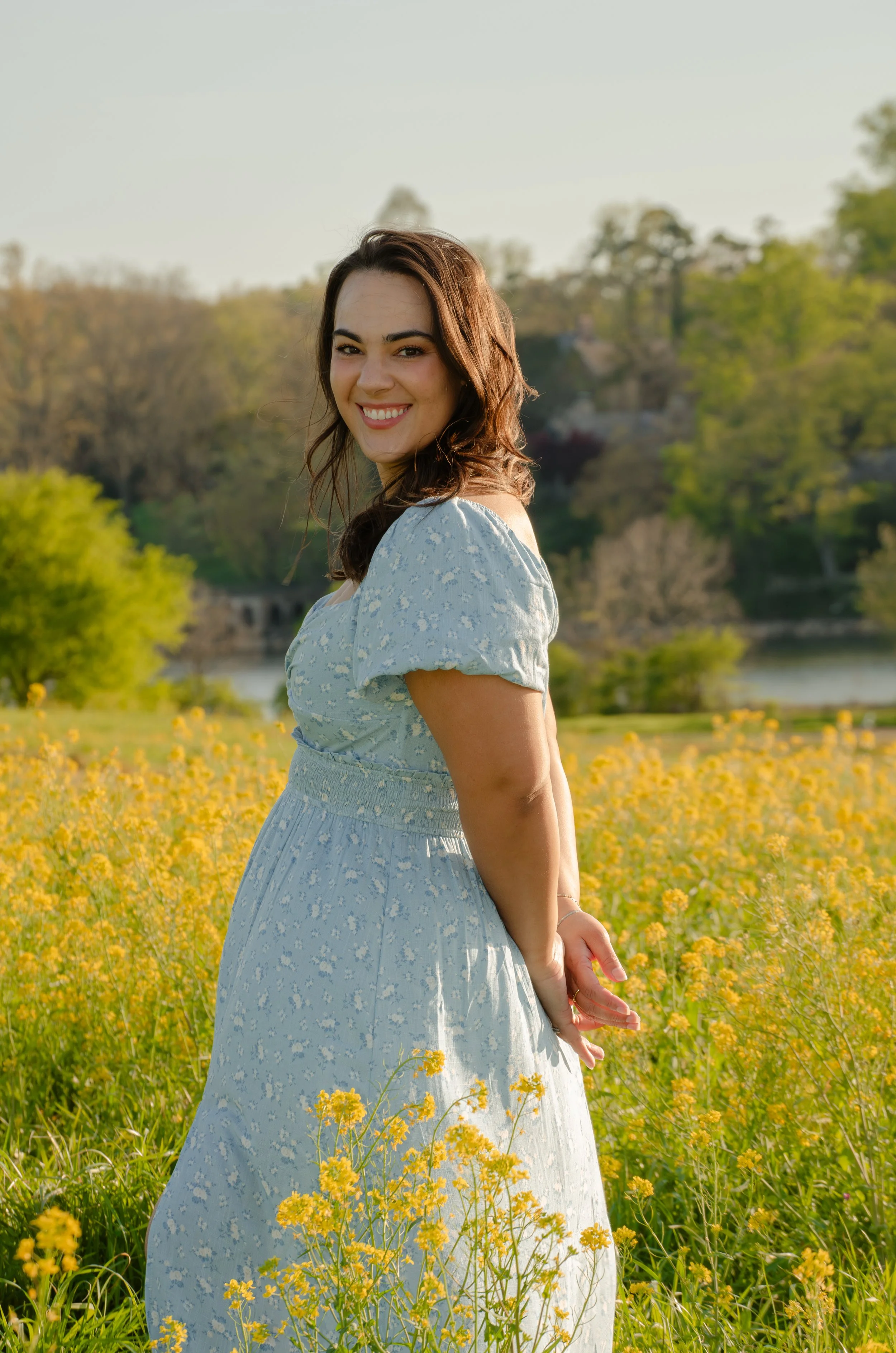 A woman in a blue floral dress smiling in a field of yellow flowers, with trees and a river in the background during daylight.