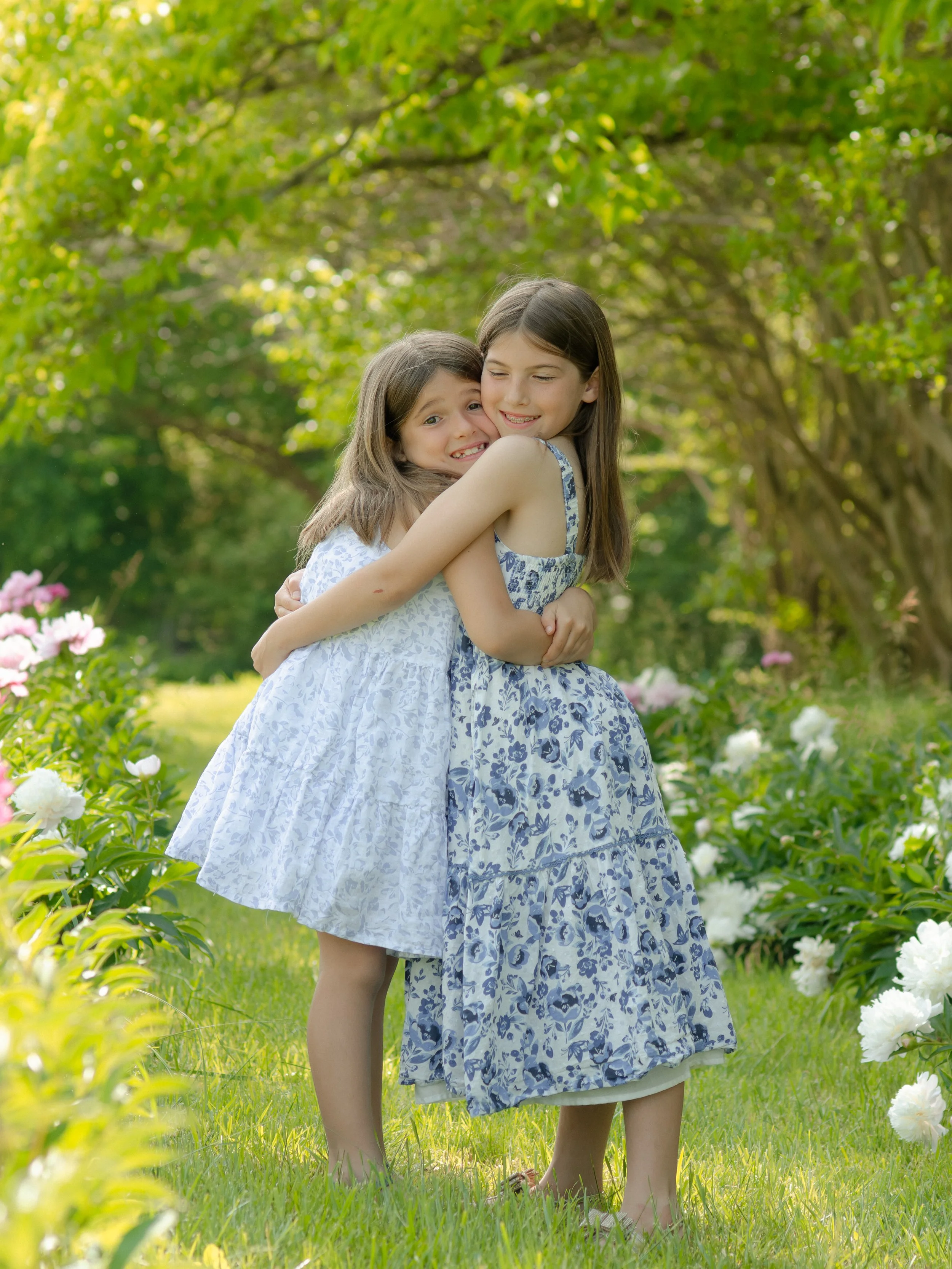 Knoxville sister photo session, hugging and smiling in a garden with trees and blooming flowers around them on a sunny day.