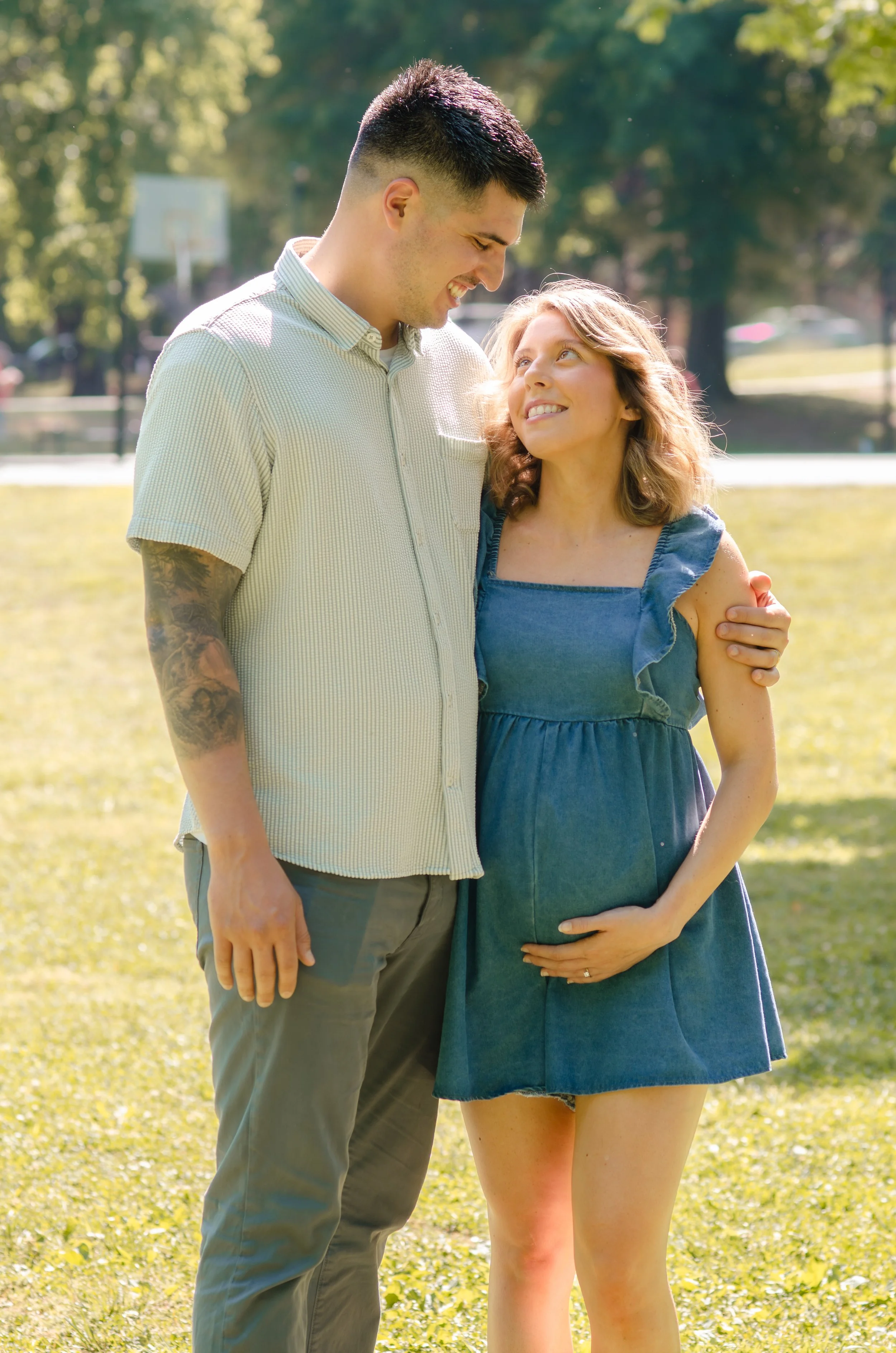 A young man and a pregnant woman stand close together on a sunny day in a park, smiling and looking at each other during a maternity photo shoot in Maryville Tn