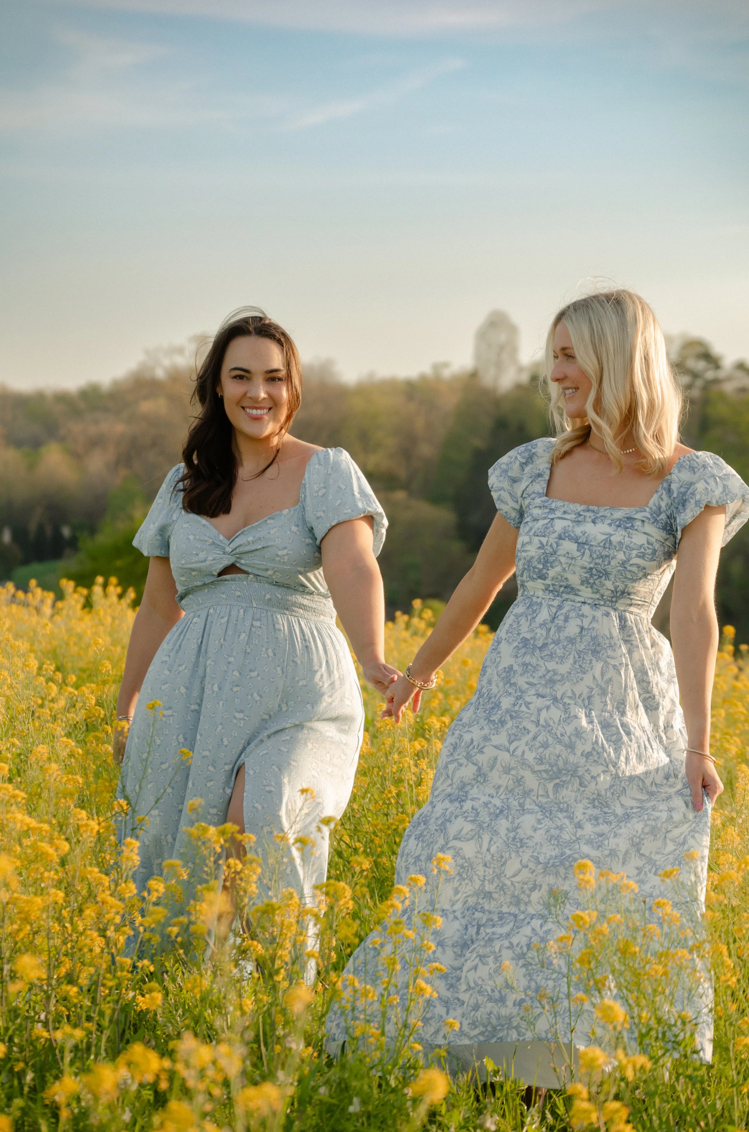 Two women holding hands and walking through a yellow flower field on a sunny day, wearing light-colored dresses with floral patterns.