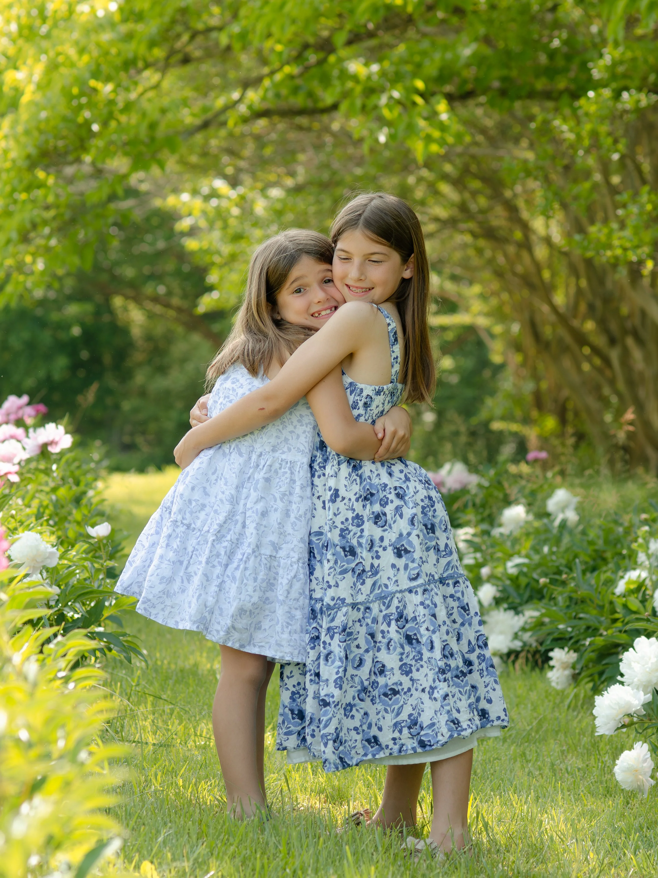 Knoxville sister photo session, hugging and smiling in a garden with trees and blooming flowers around them on a sunny day.