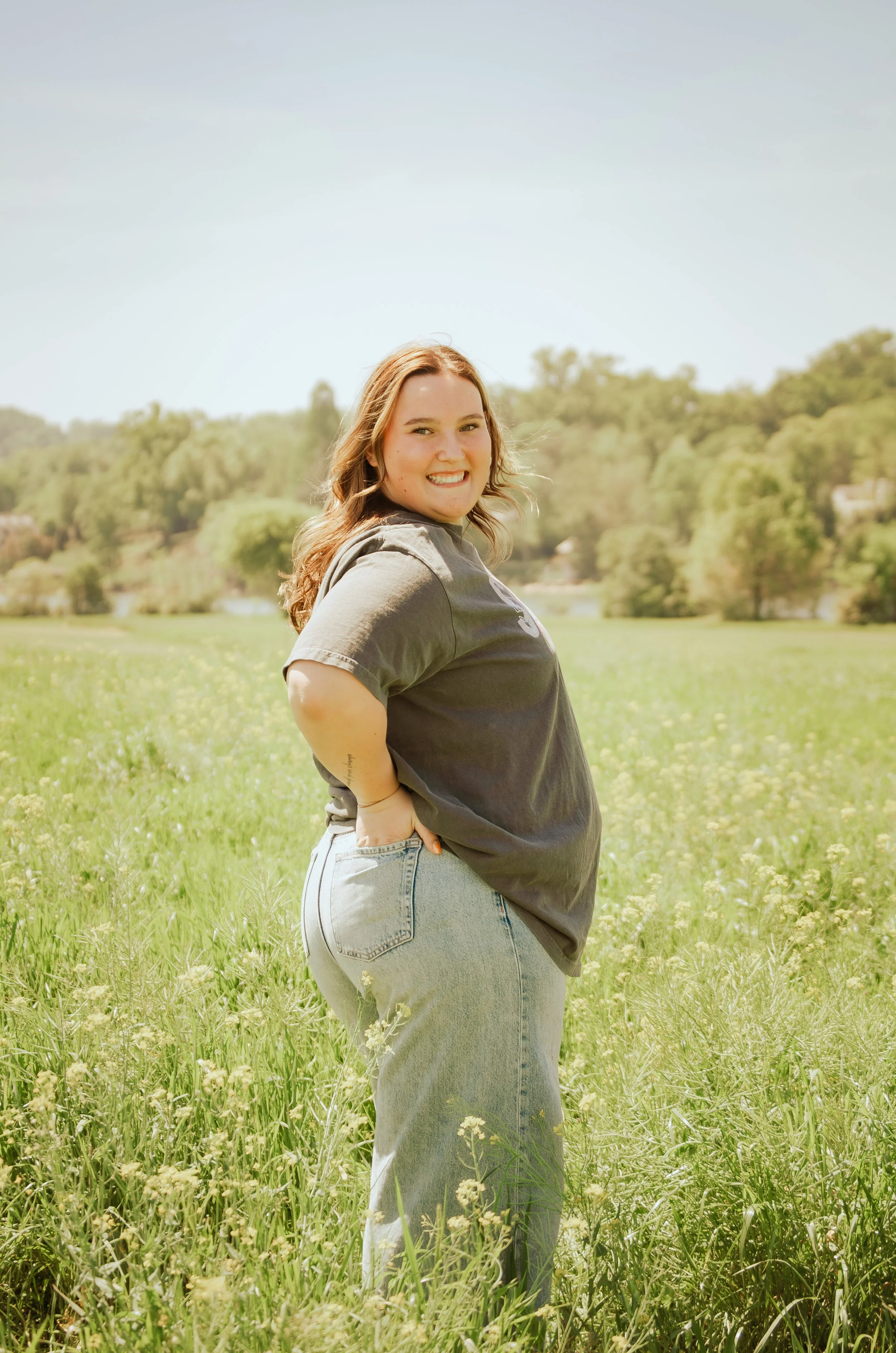 Young woman with long hair smiling in a green field with trees and hills in the background.