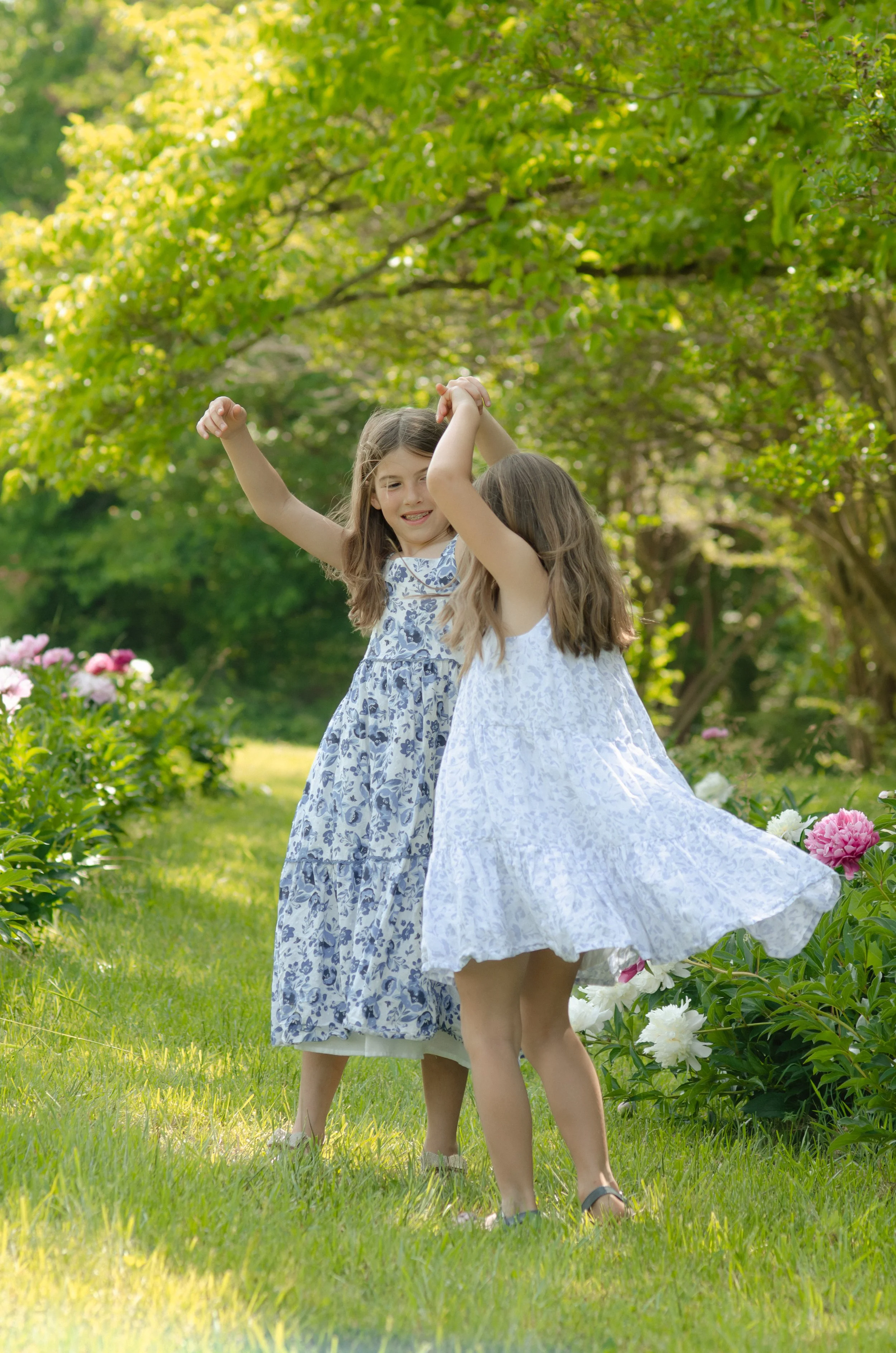 Knoxville photosession with two sisters playing and dancing together outdoors in a beautiful garden with flowering bushes and trees on a sunny day.