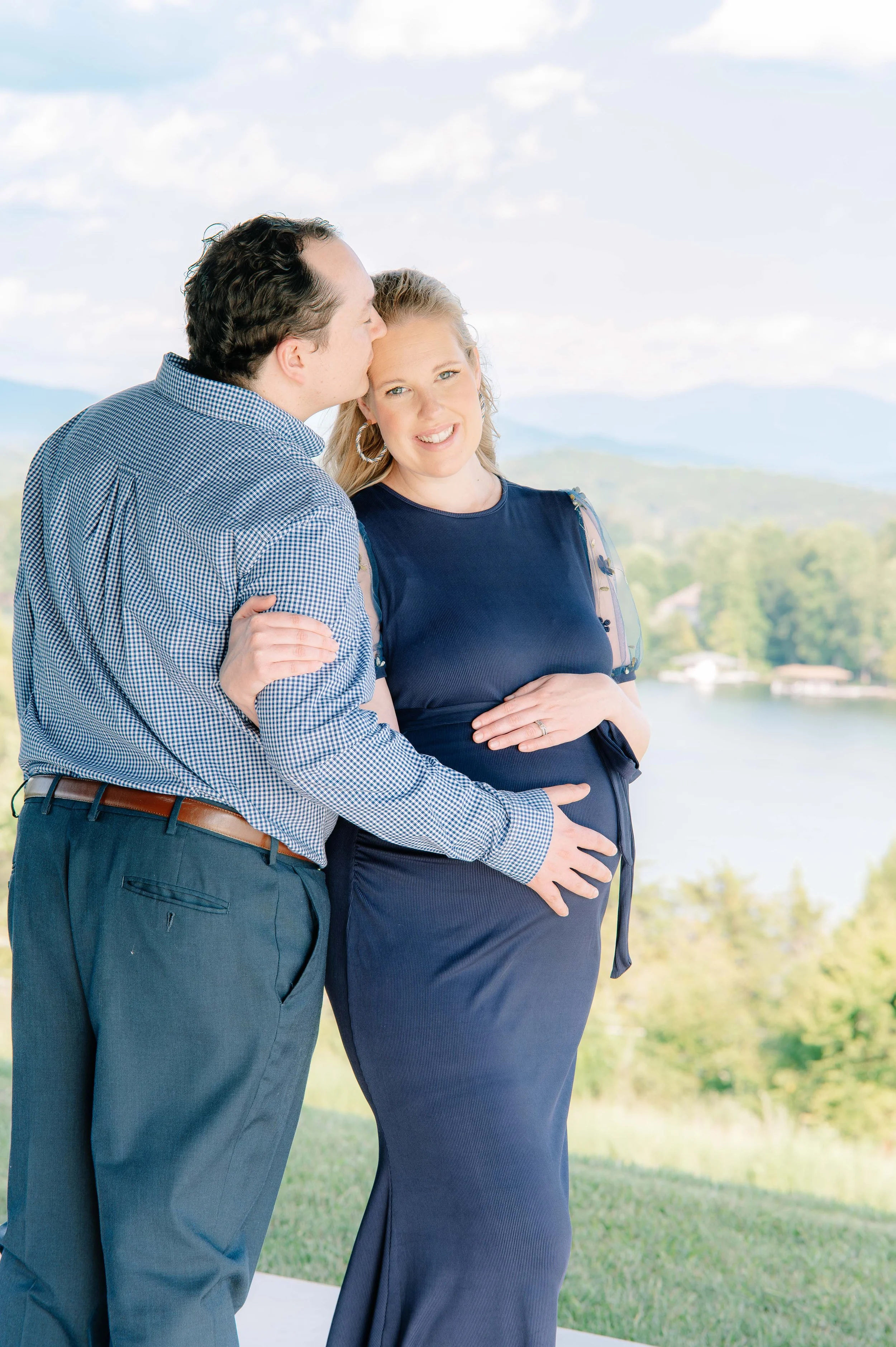 Husband and wife smiling together in a Vonore, Tennessee field with mountain and lake views during their maternity session