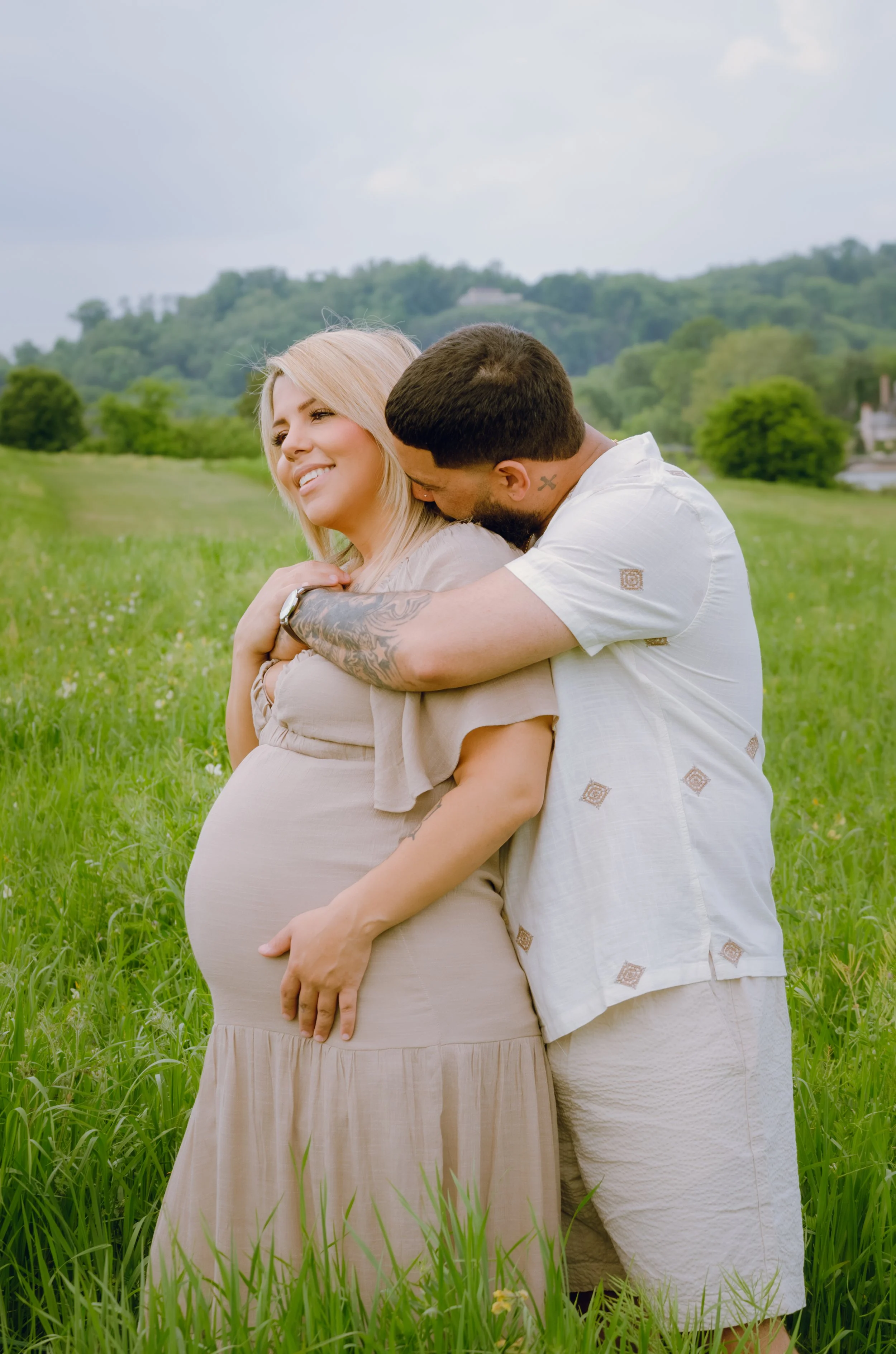 A pregnant woman with blonde hair smiling as she stands in a field of tall grass, while a man with dark hair and a beard hugs her from behind, kissing her on the head, with a green hilly landscape and cloudy sky in the background.