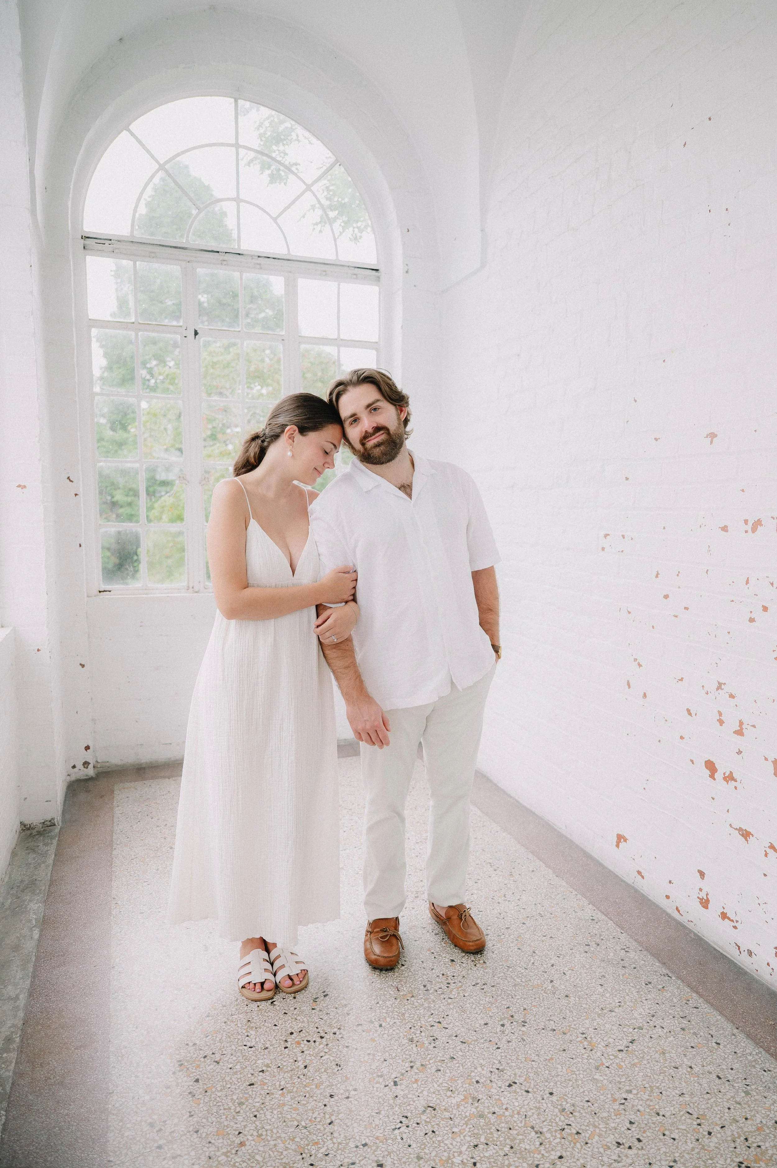 A young woman and man standing inside a bright, white brick building with large arched windows. The woman is wearing a white dress with spaghetti straps and sandals, and the man is wearing a white shirt, white pants, and brown shoes. They are close together, with the woman's head resting on the man's shoulder and her eyes closed.