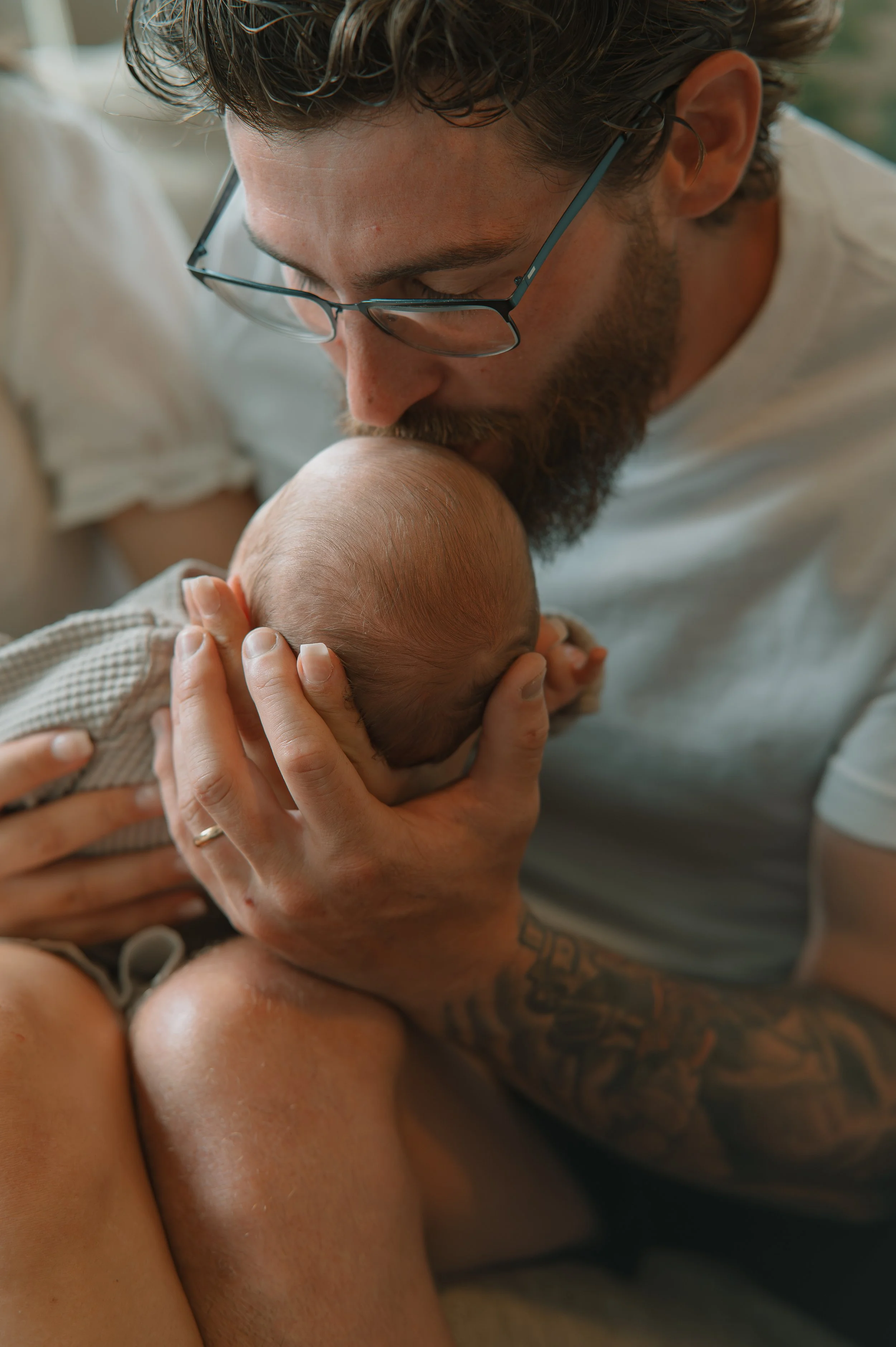A man wearing glasses is gently kissing a baby's head, holding the baby securely while sitting on an orange sofa.