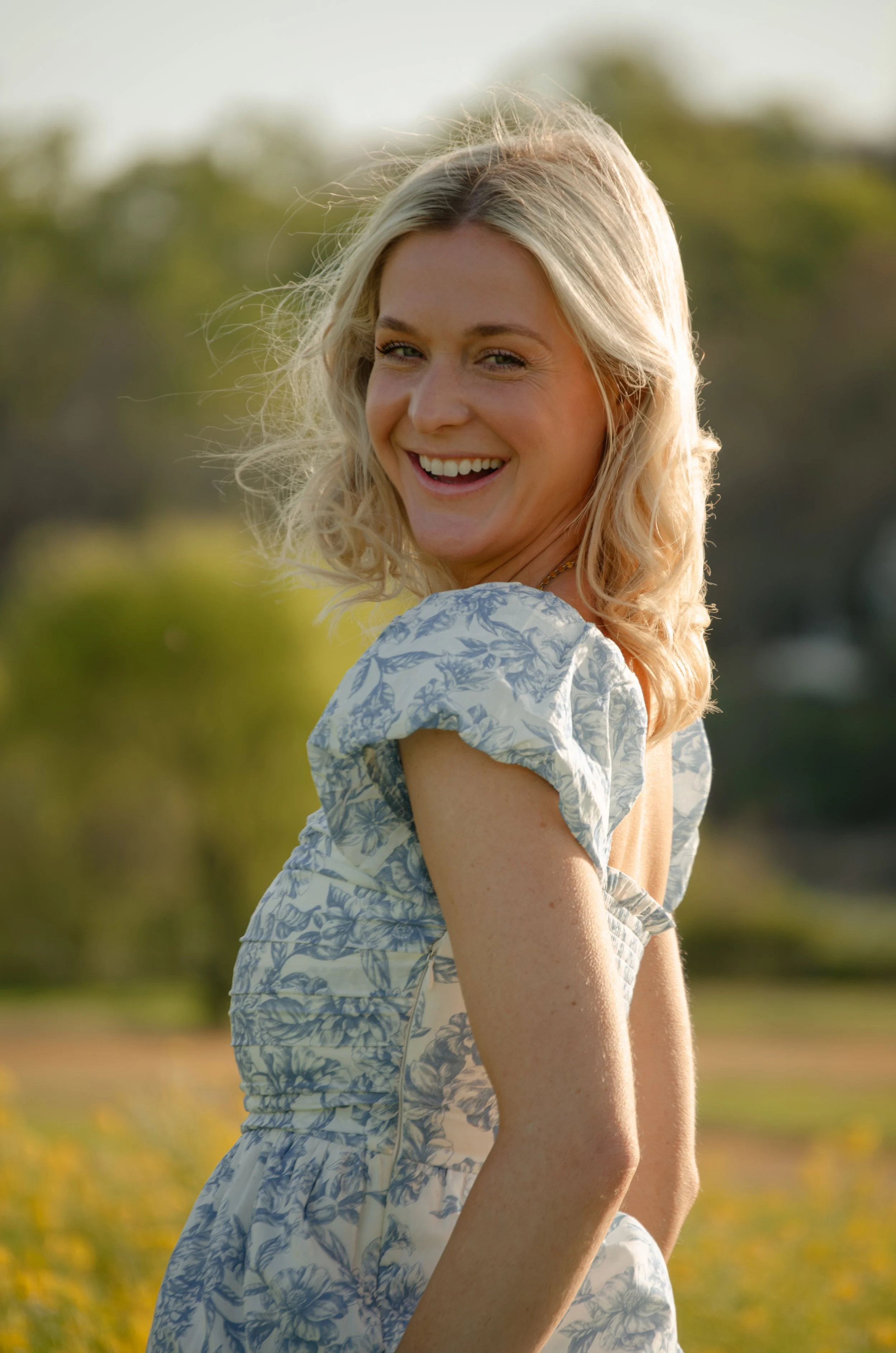 A smiling woman with blonde, curly hair in a floral dress outdoors on a sunny day, with blurred greenery in the background.