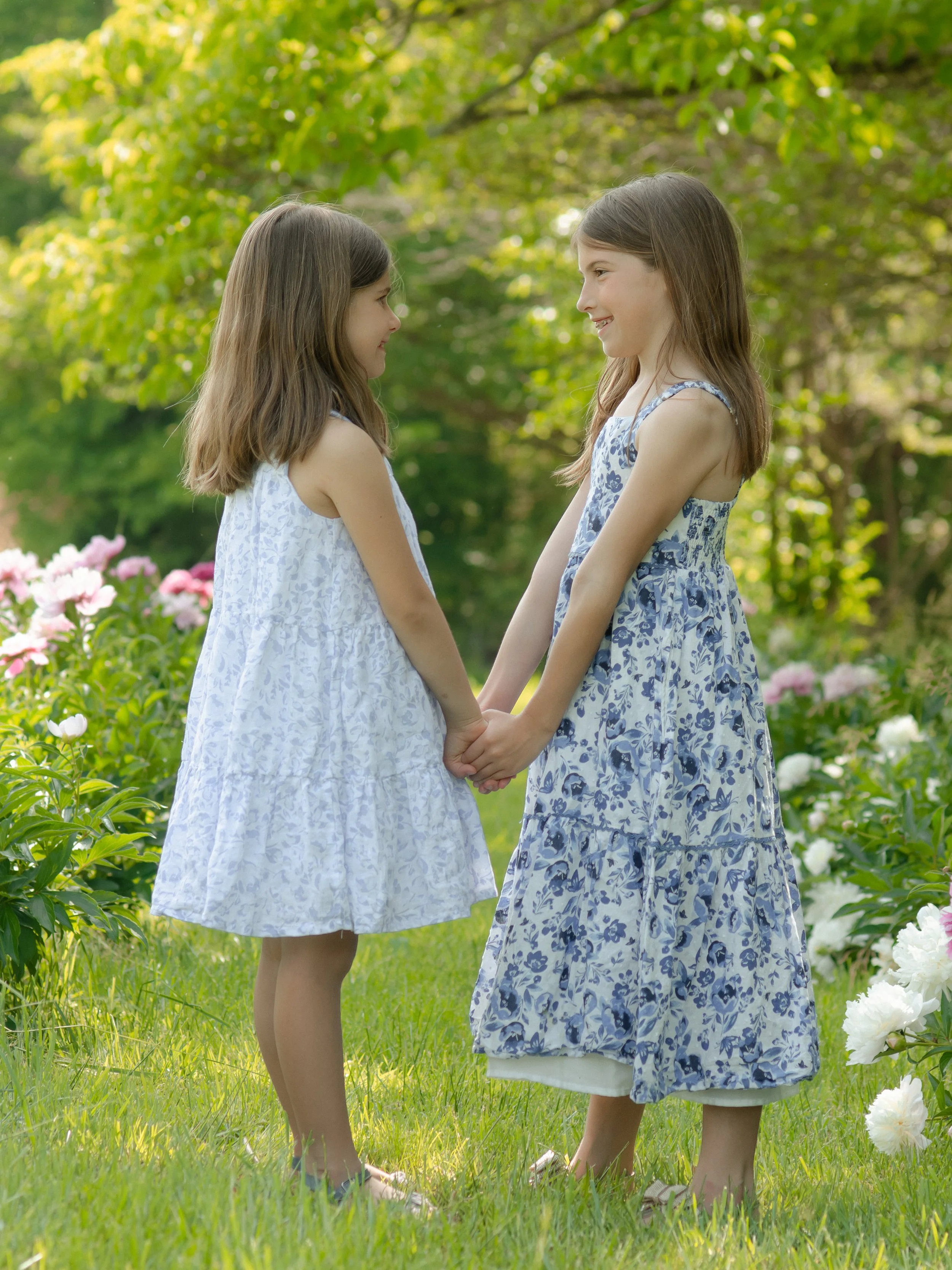 Knoxville photosession with two sisters holding hands and facing each other in a garden with trees and flowers, dressed in light blue and white floral dresses.