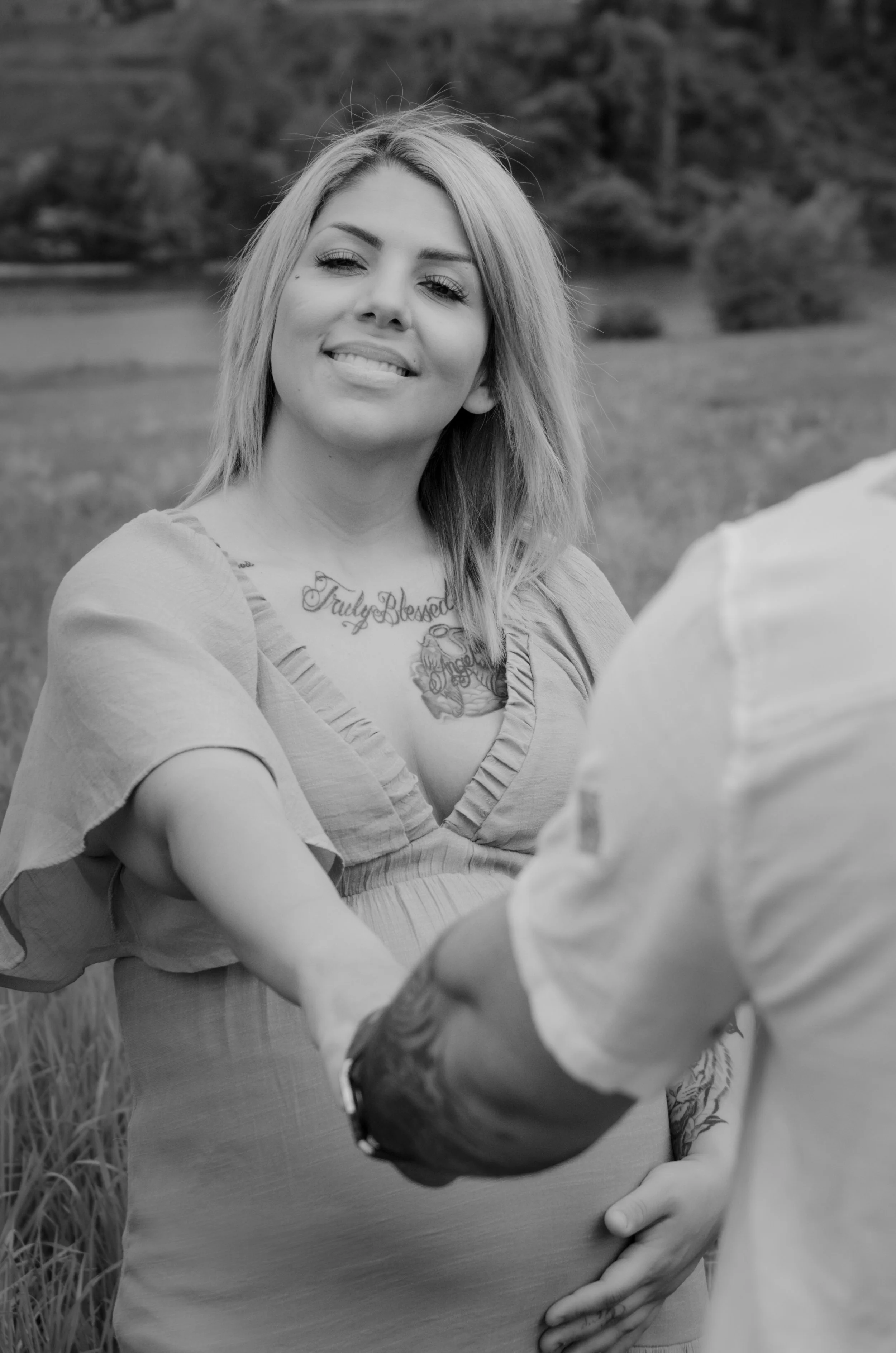 A woman with light-colored hair and tattoos on her chest smiling, standing outdoors in a natural setting, interacting with a person in a light-colored shirt.