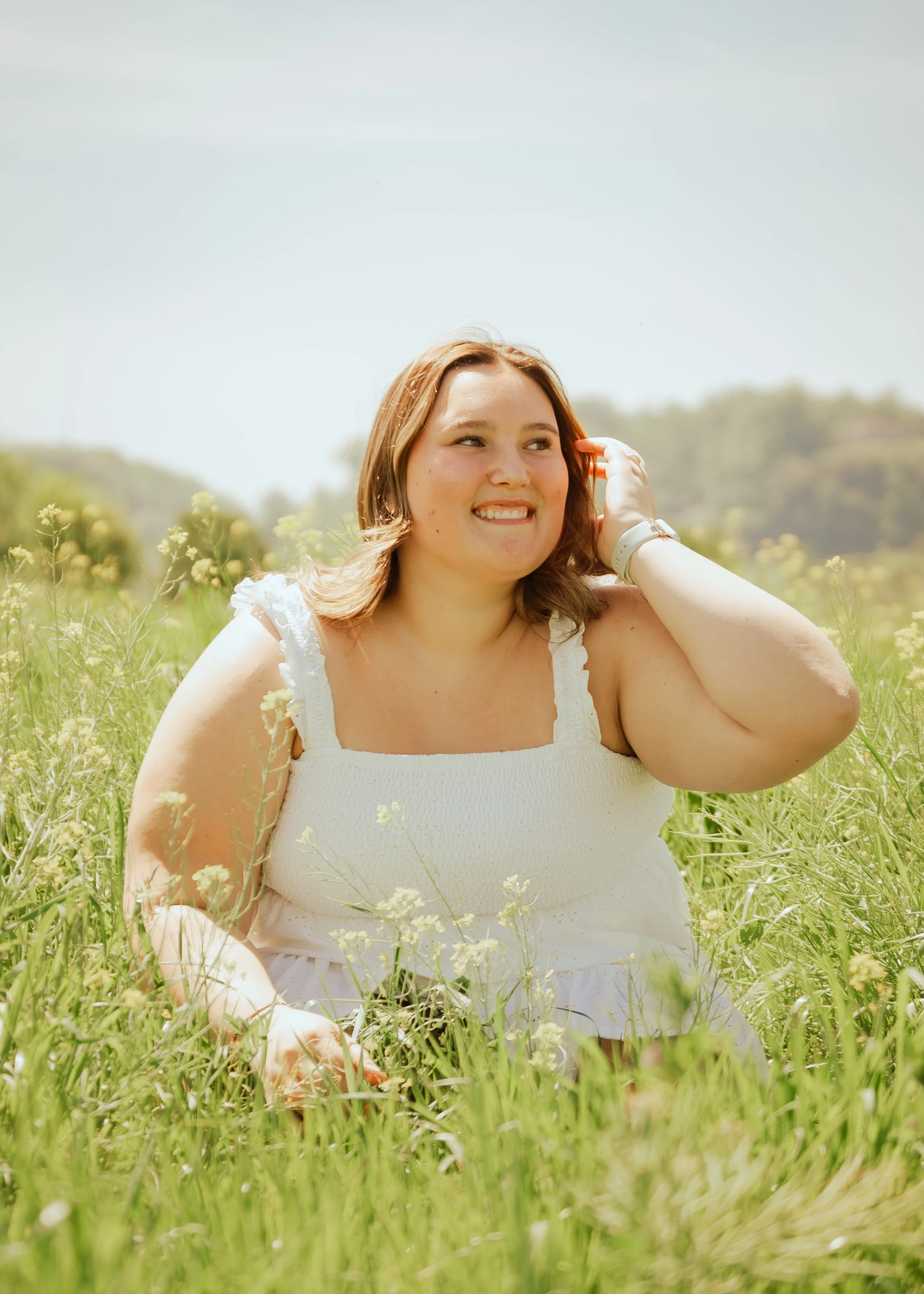 A smiling woman with light brown hair, wearing a white dress, sitting in a field of green grass and yellow flowers, with hills in the background and a clear blue sky.