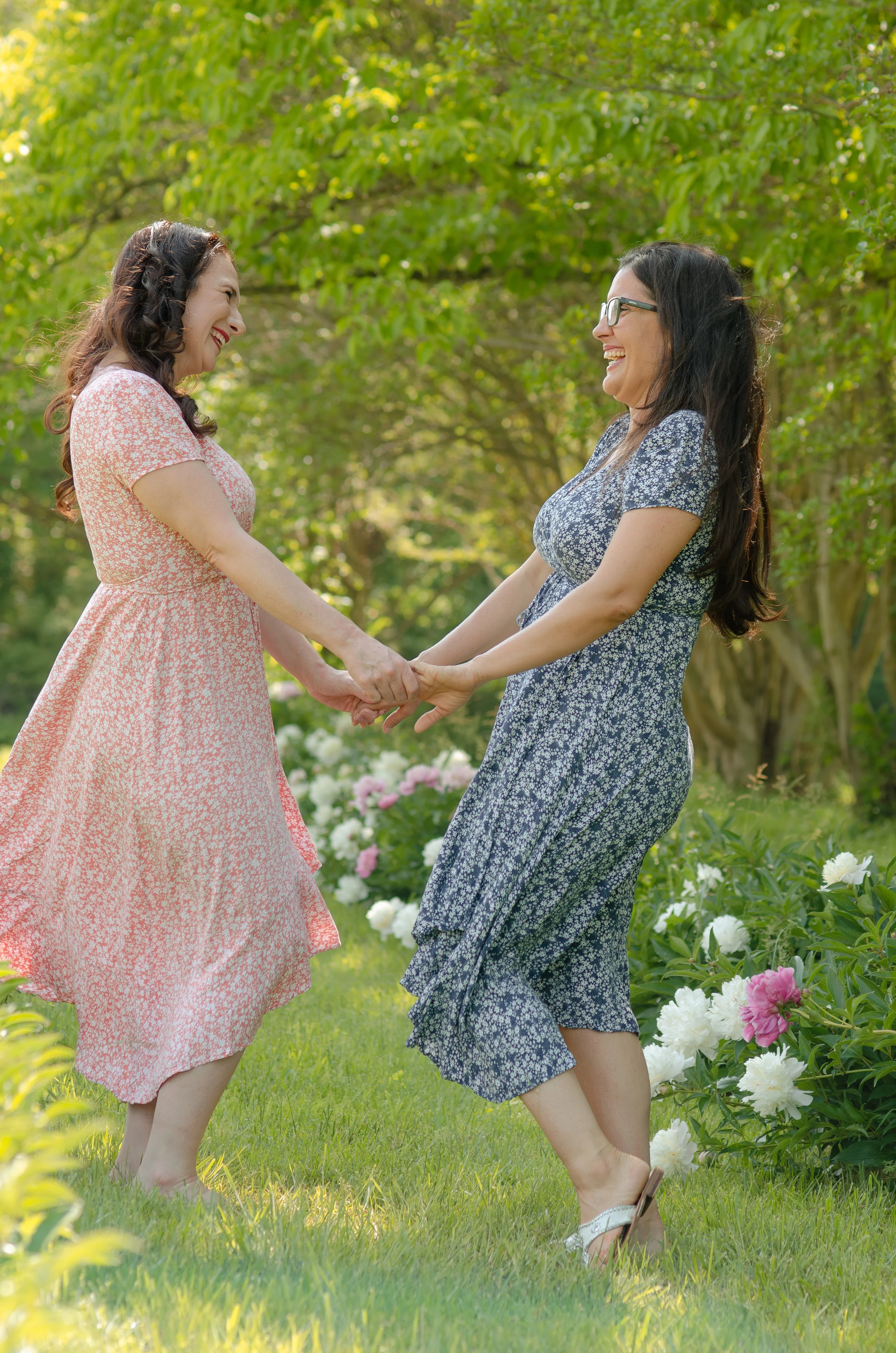Photo session with adult sisters holding hands and smiling at each other outdoors in a garden with blooming peonies and green trees.