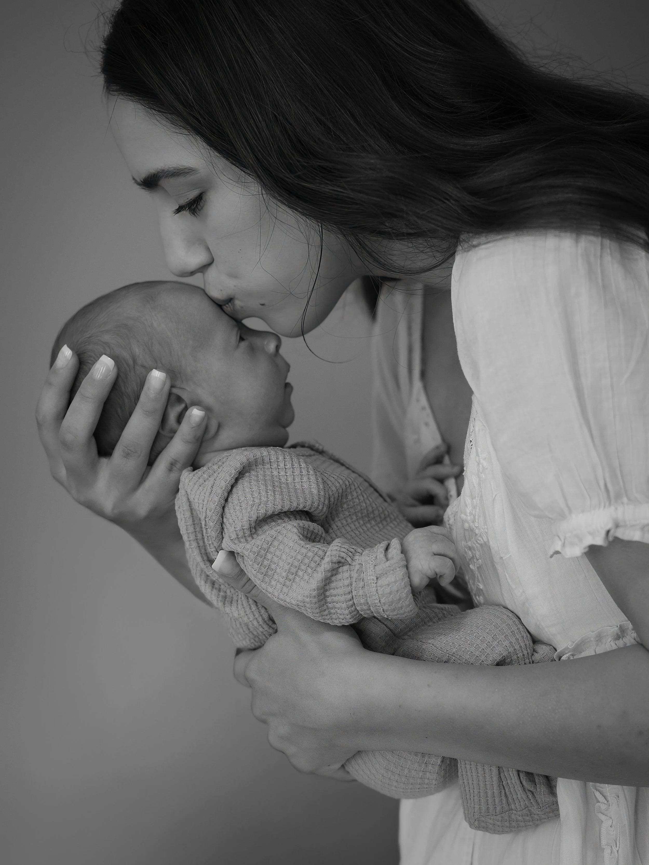 A woman with long dark hair is gently kissing a baby on the forehead, holding the baby's head with one hand. The baby looks up at the woman, and they are both dressed in light-colored clothing. The background is plain and blurred.