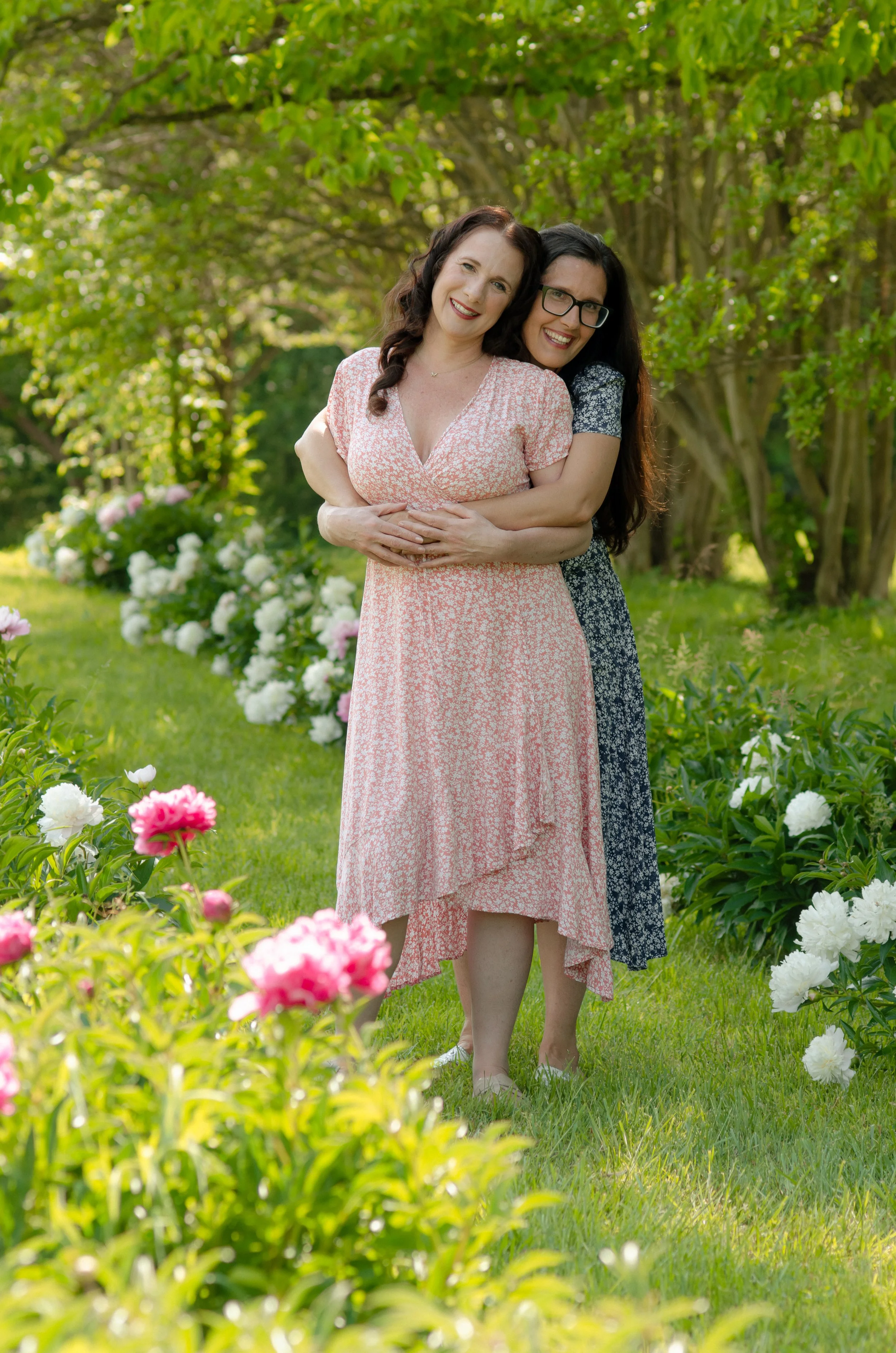 Knoxville adult sister session with loving sisters in natural outdoor environment, adult sisters smiling and hugging each other in a garden with blooming flowers and green trees.