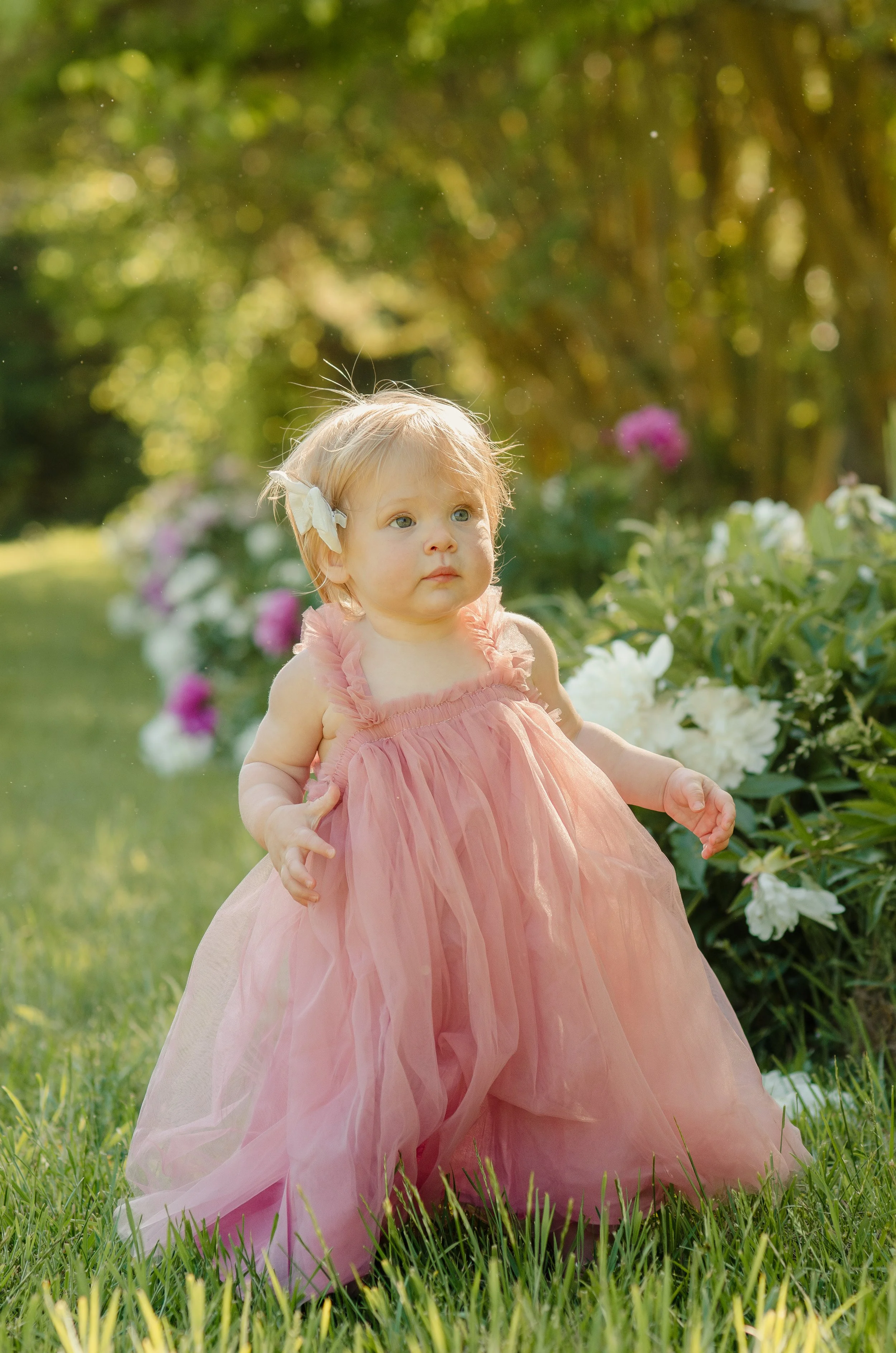 A young girl in a pink dress standing in a garden with green grass and colorful flowers, with trees and sunlight in the background.