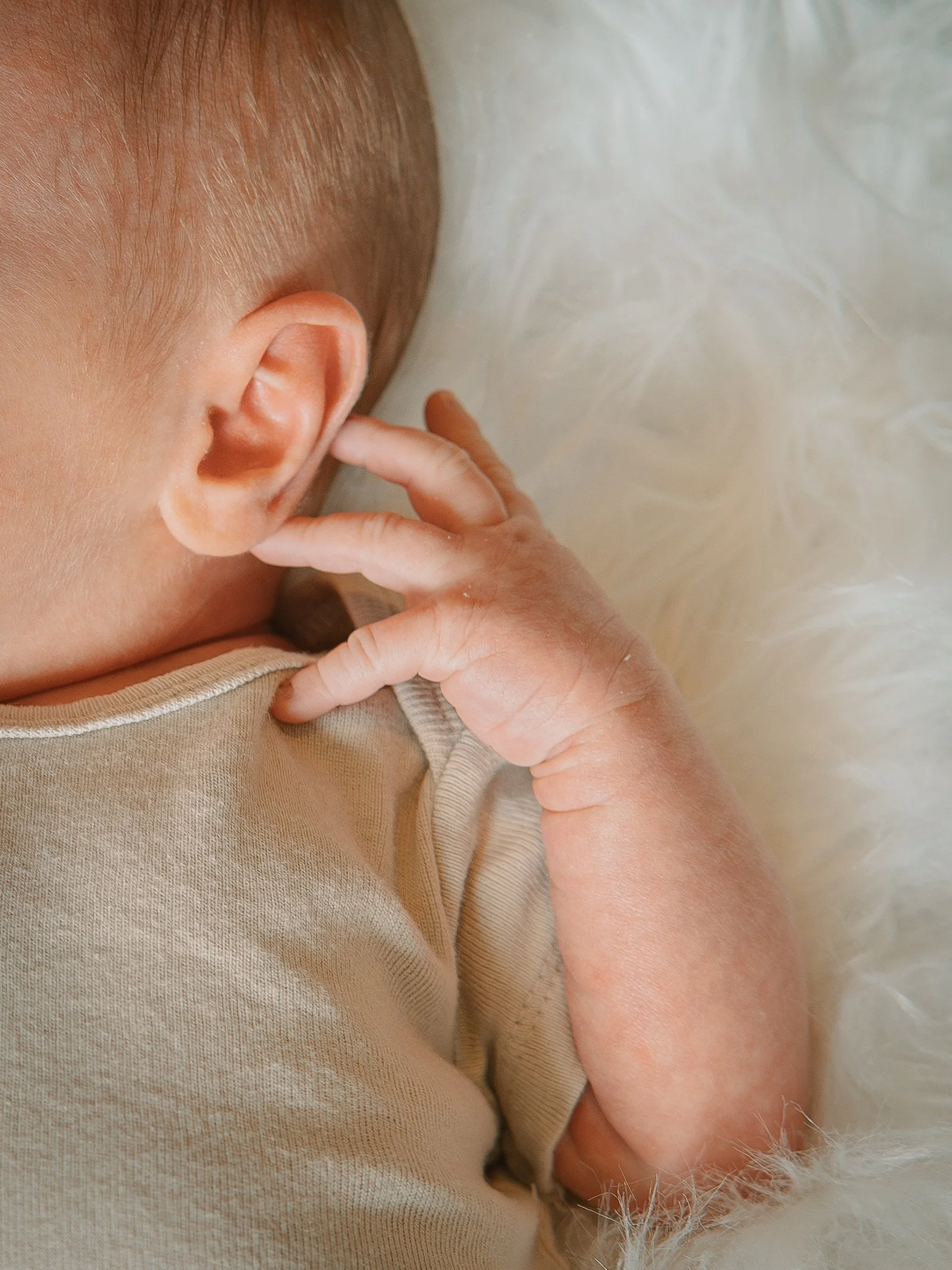 Close-up of a baby with their finger in their ear, lying on a soft, cream-colored surface
