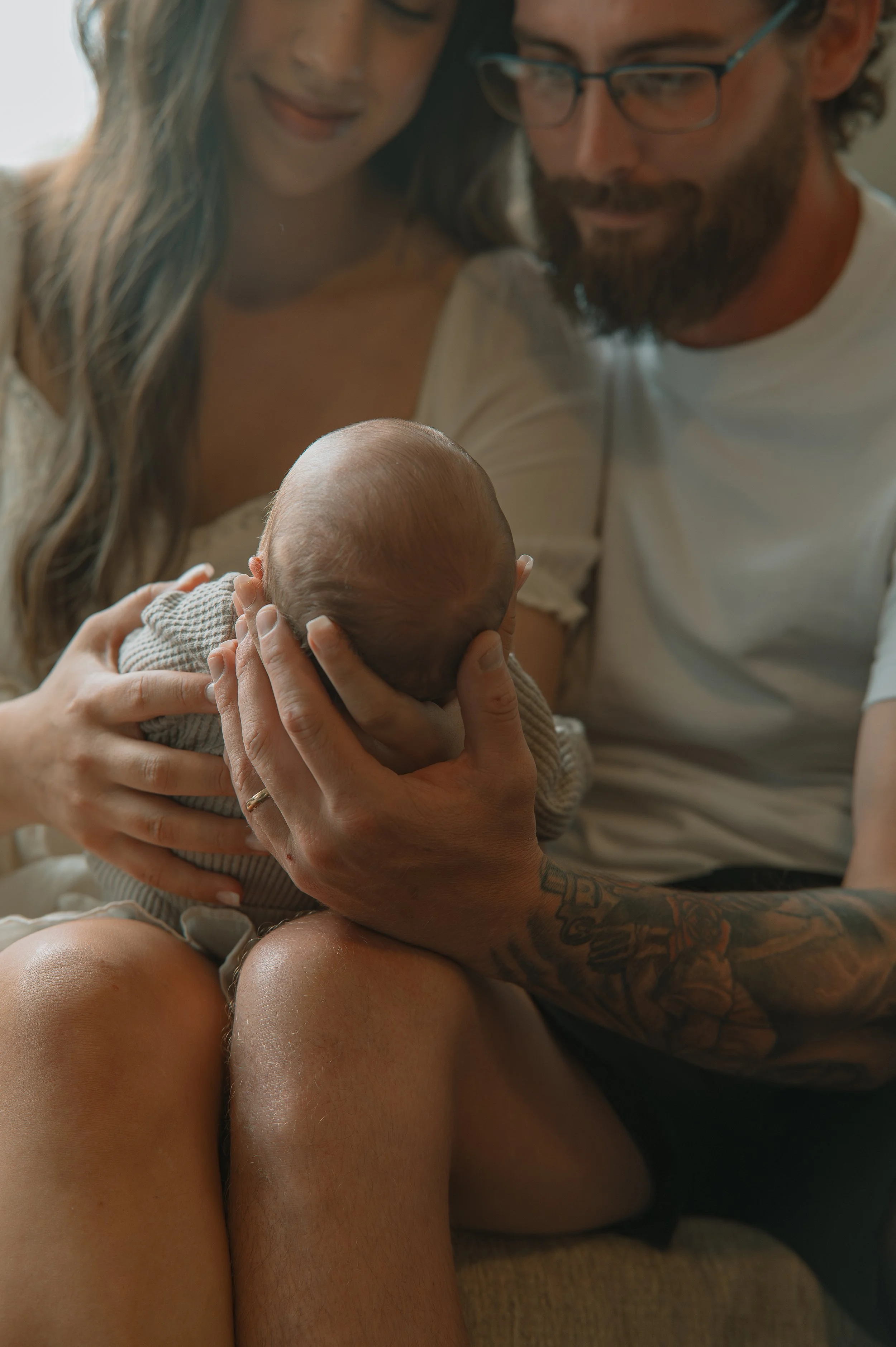 A woman and man look at a baby they are holding. The woman has long hair and the man has a beard, glasses, and tattoos. The baby looks like an infant with a bald head and is cozy in their arms.