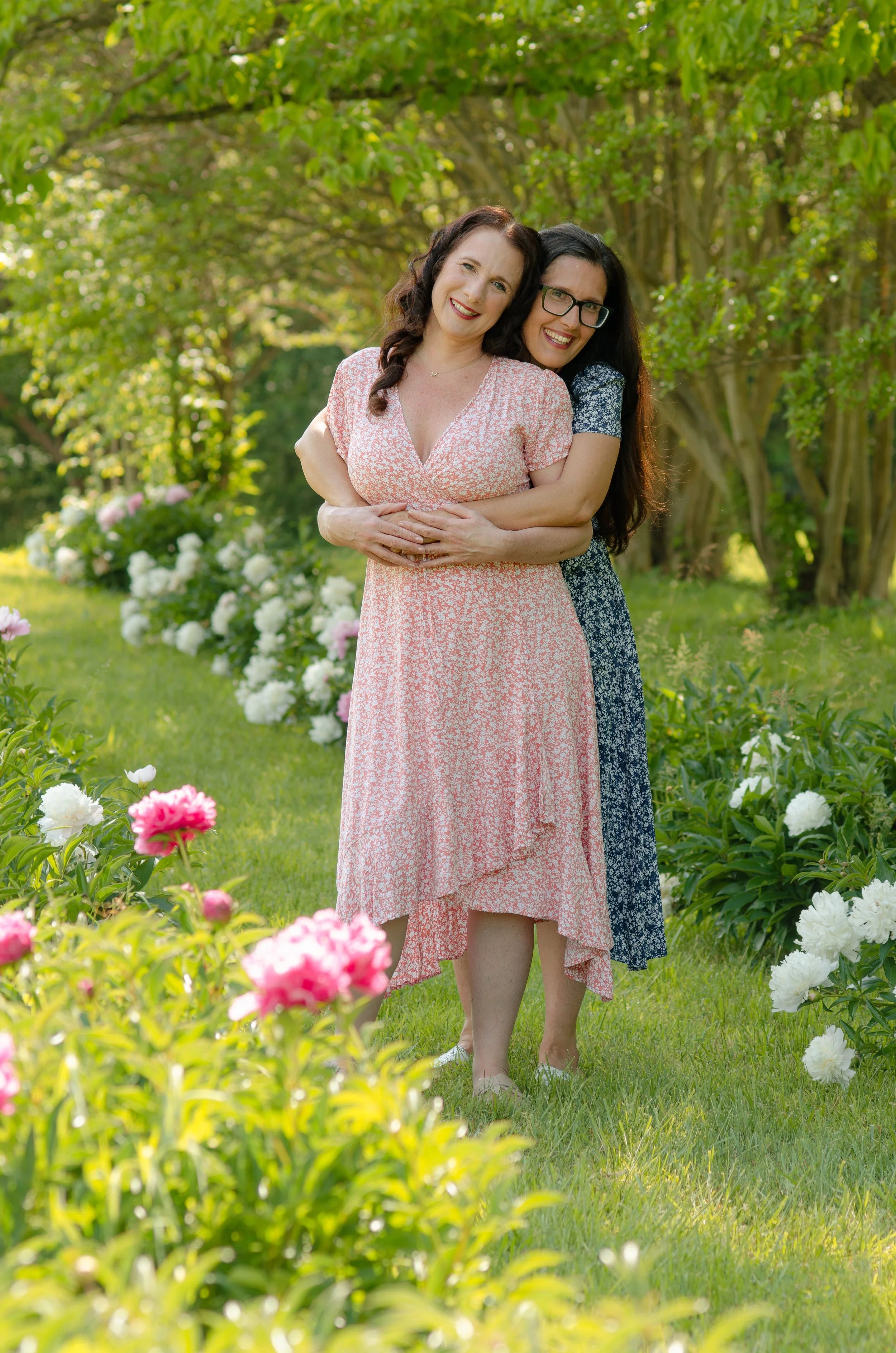 Knoxville adult sister session with loving sisters in natural outdoor environment, adult sisters smiling and hugging each other in a garden with blooming flowers and green trees.