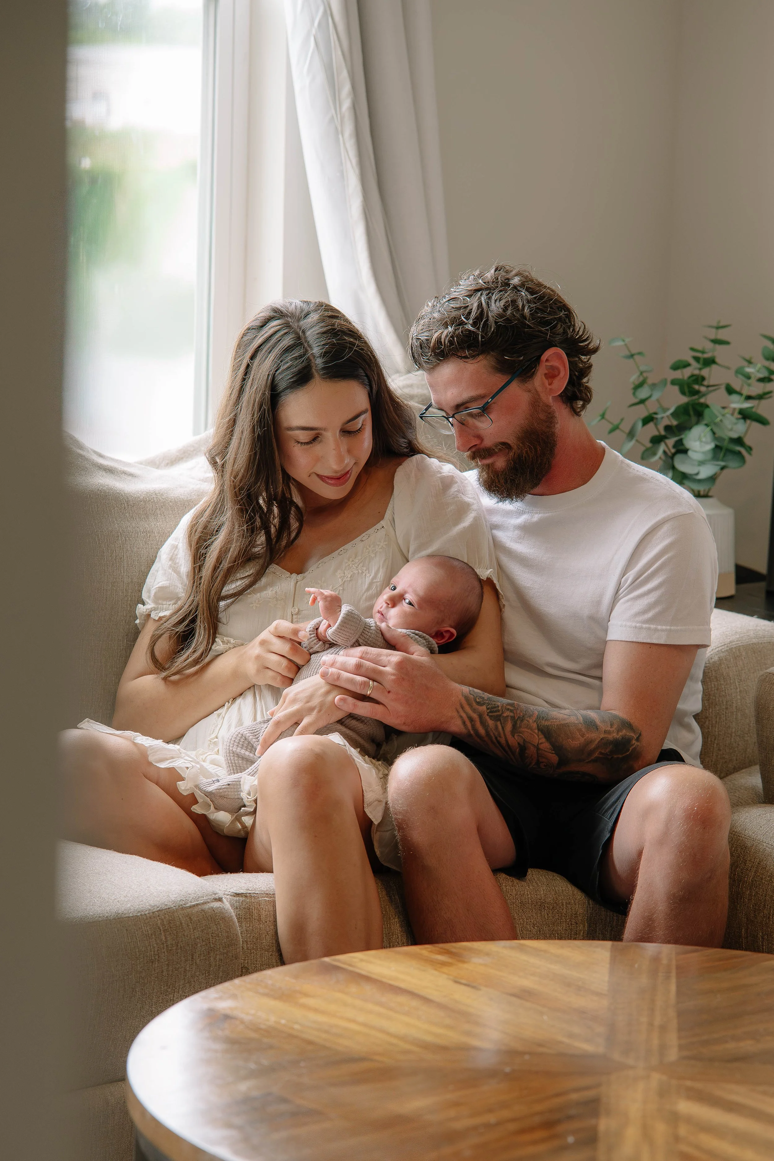 A couple with a baby sitting on a beige couch in a bright living room, near a window with curtains, with a wooden coffee table in the foreground.
