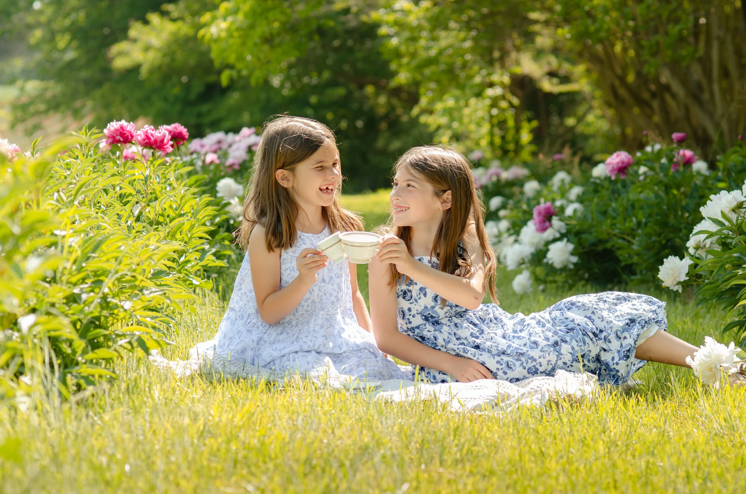 Two sisters in a photo session sitting on a white blanket in a garden with colorful flowers, holding teacups, smiling and enjoying a sunny day.