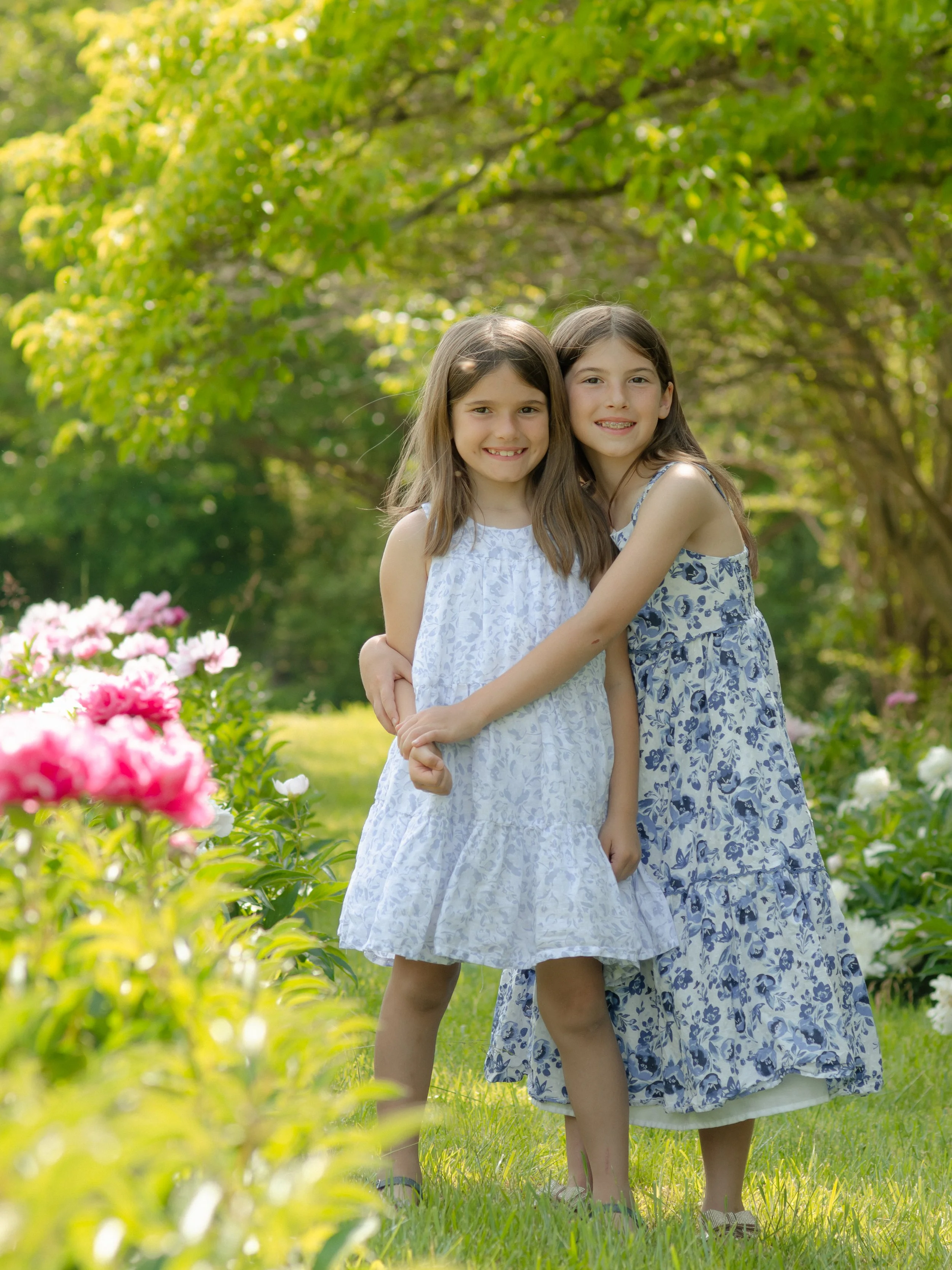 Two sisters in dresses standing and hugging each other in a garden with blooming flowers and green trees in the background.