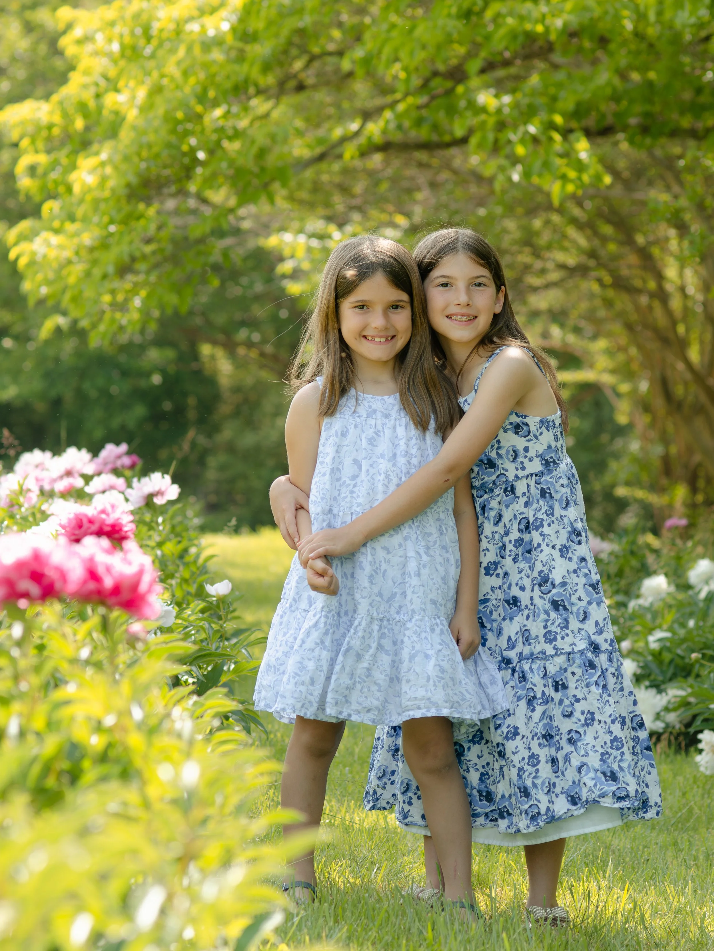 Two sisters in dresses standing and hugging each other in a garden with blooming flowers and green trees in the background.