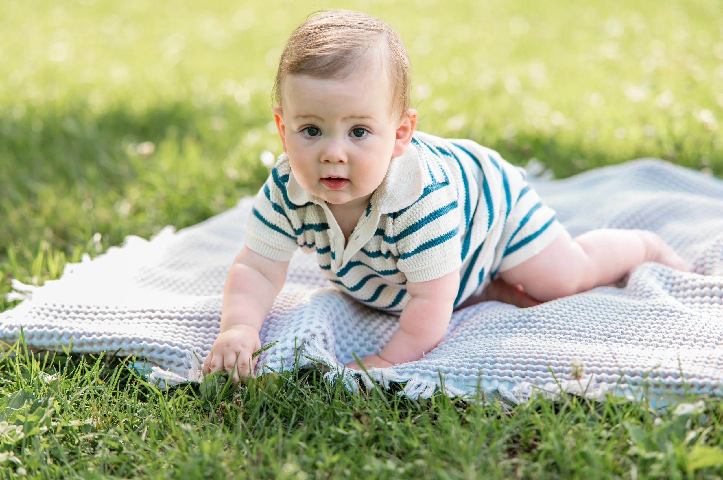 A young baby crawling on a blanket on the grass outdoors with green trees and sunlight in the background.