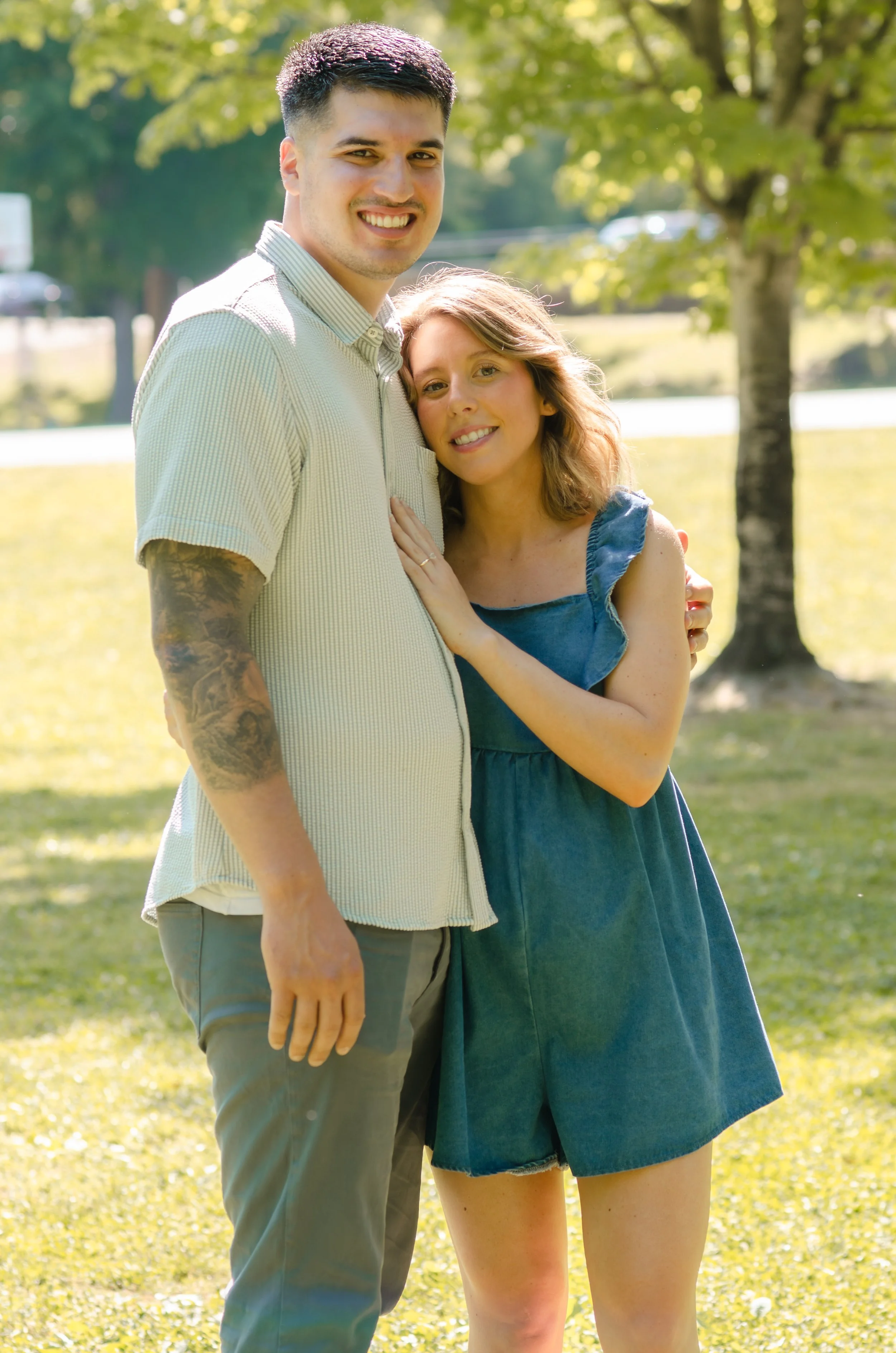 A young couple standing outdoors on a sunny day, smiling, with trees and a grassy area in the background at Sandy Springs in East Tennessee, during a maternity photography session