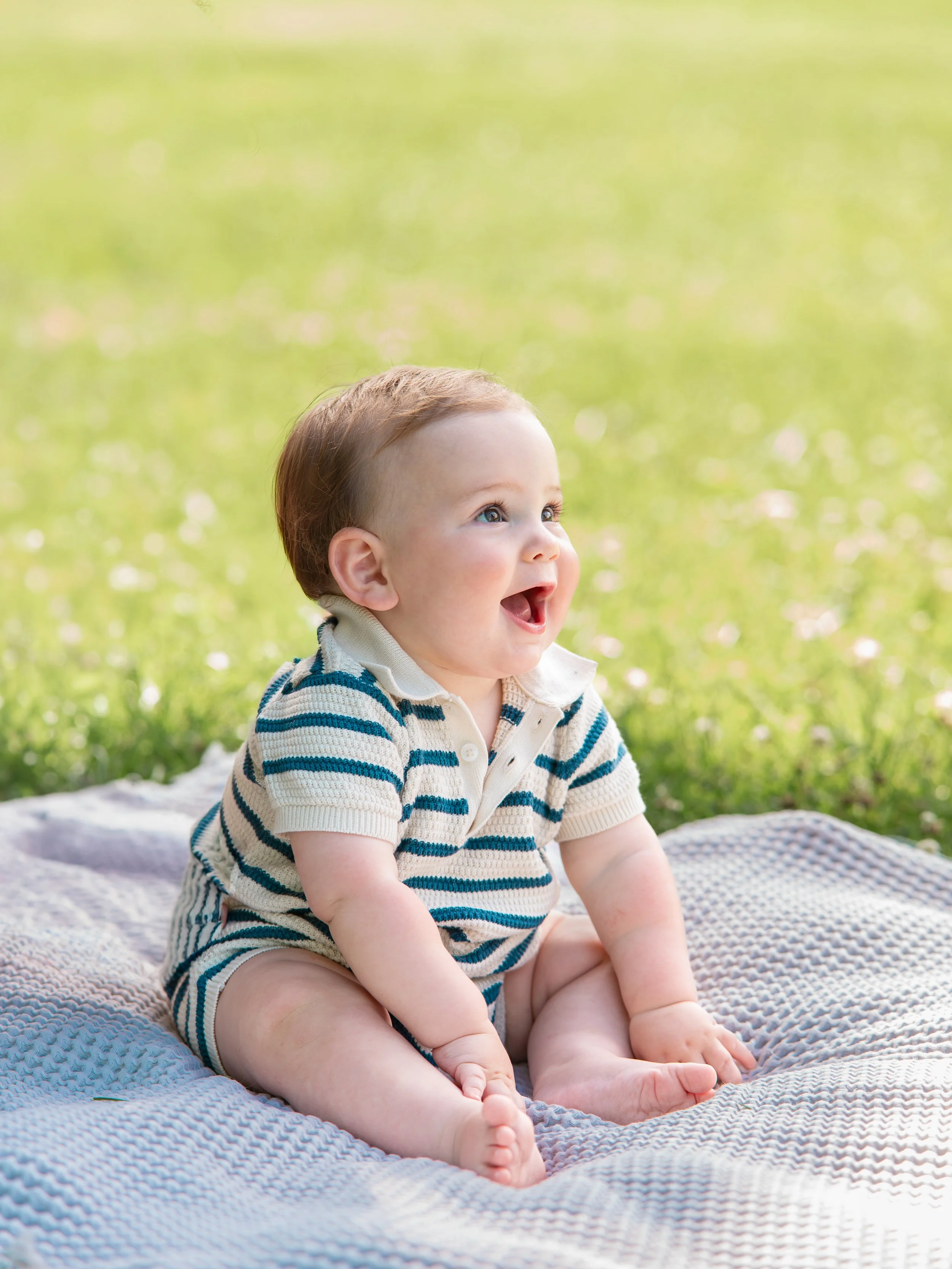 Young baby with brown hair and blue eyes sitting on a blanket outdoors with a green grassy background, wearing a striped beige and blue shirt, looking happy and excited.