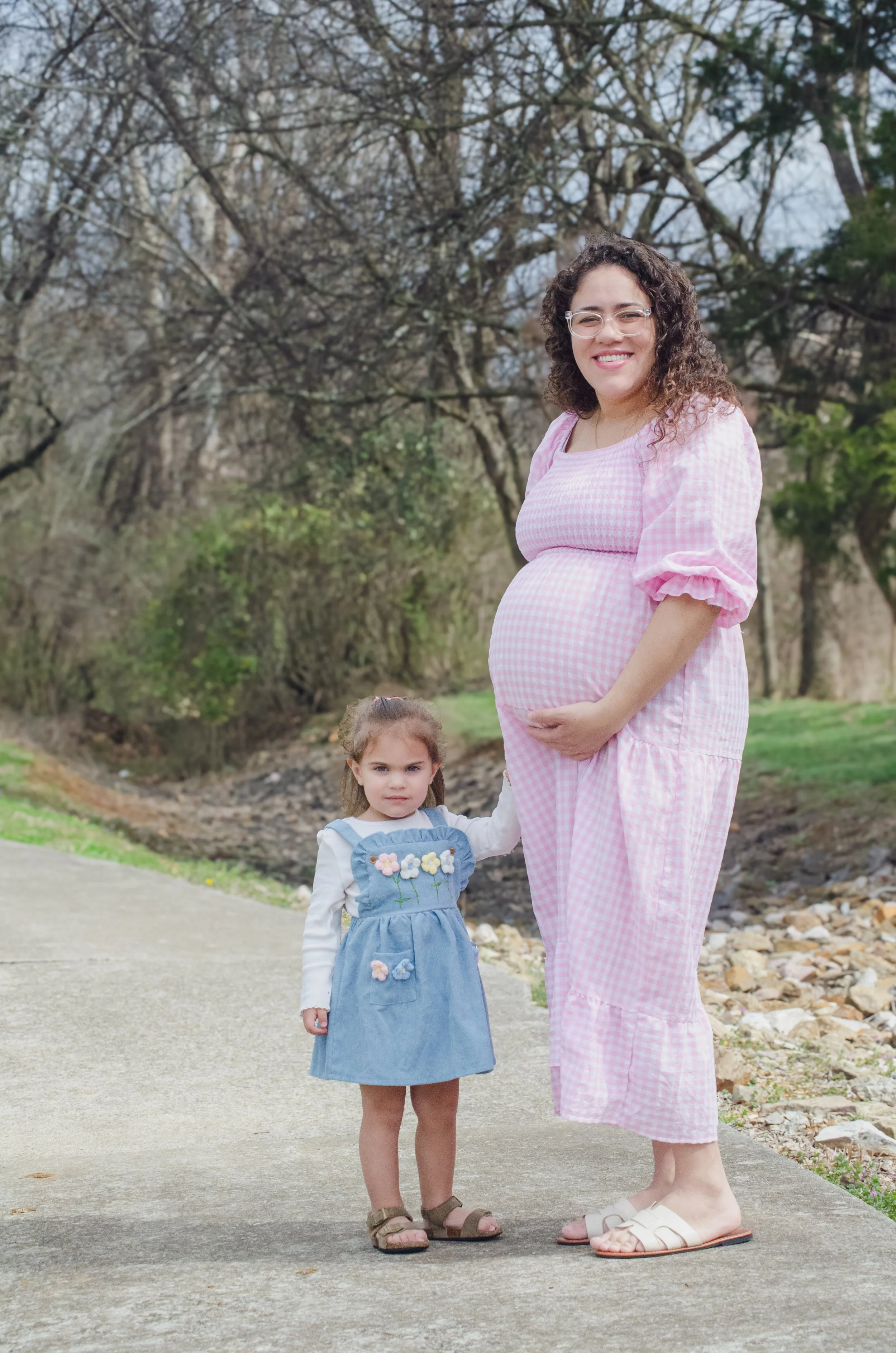 A pregnant woman smiling and standing outdoors with a young girl, both wearing casual clothing, on a pathway near trees and rocks.
