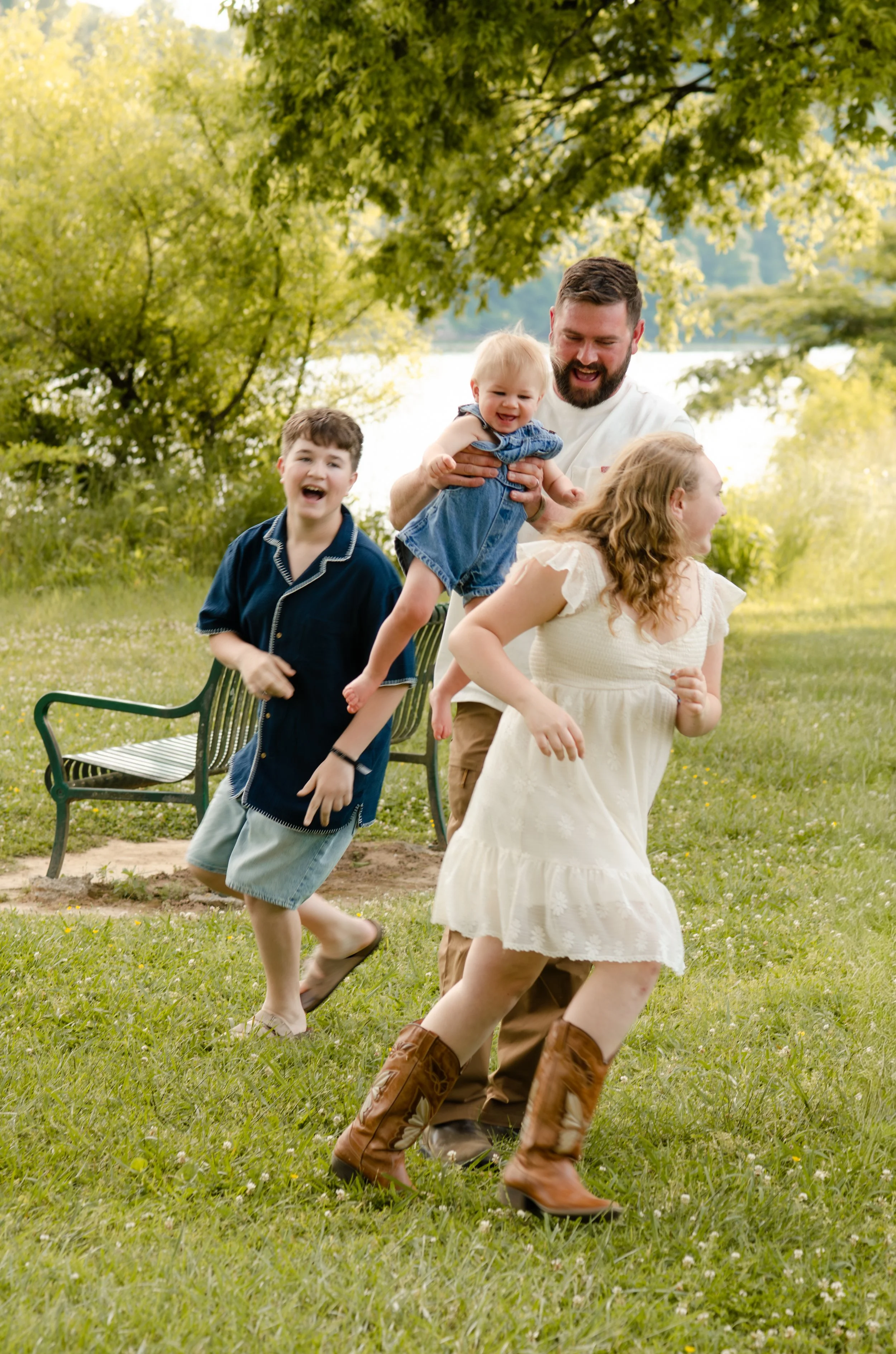 A family of five outdoors near a lake, playing and laughing on the grass with trees and water in the background during a family photoshoot
