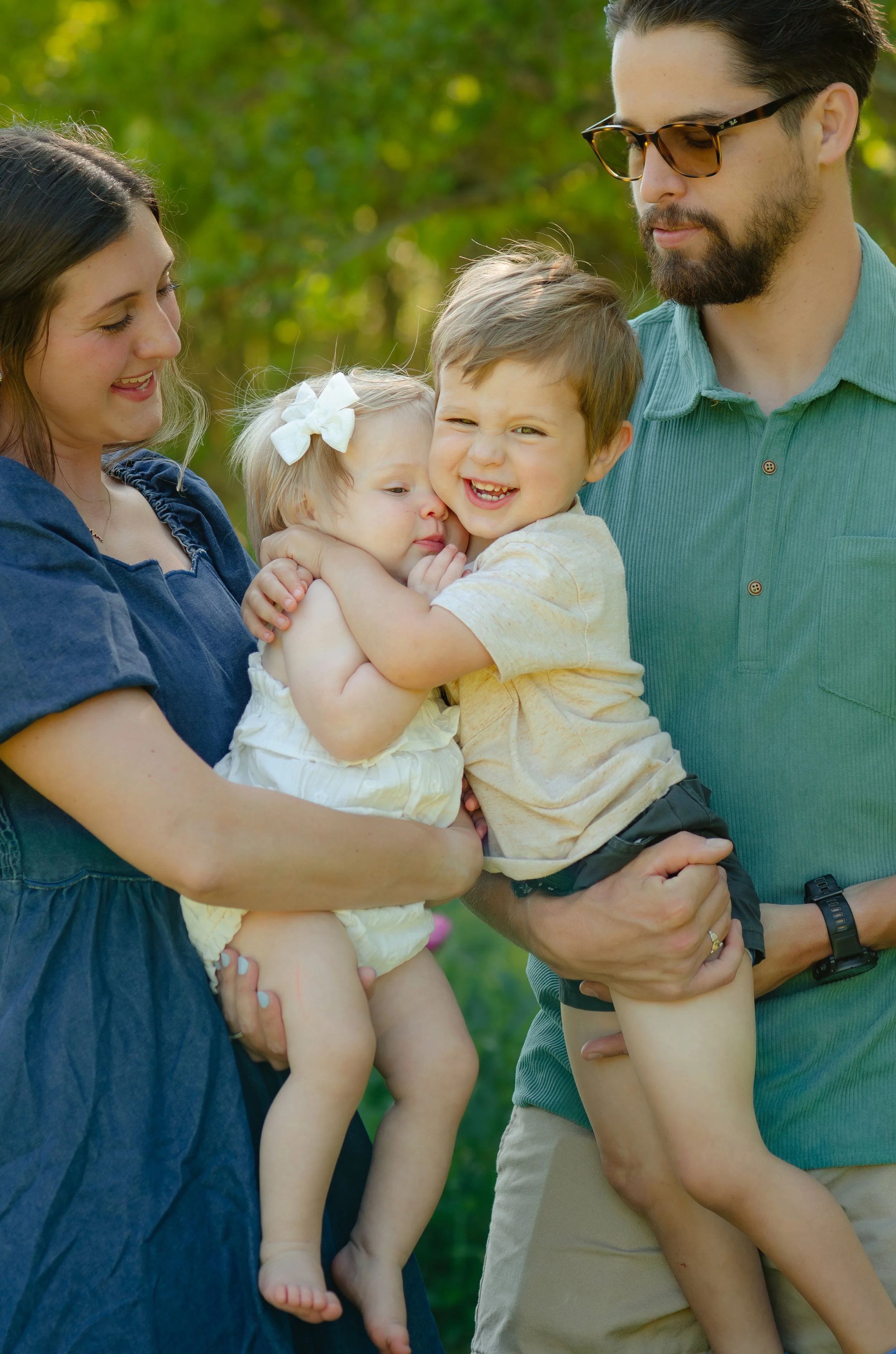 A family of five outdoors in a park, smiling and hugging each other, with green trees in the background.