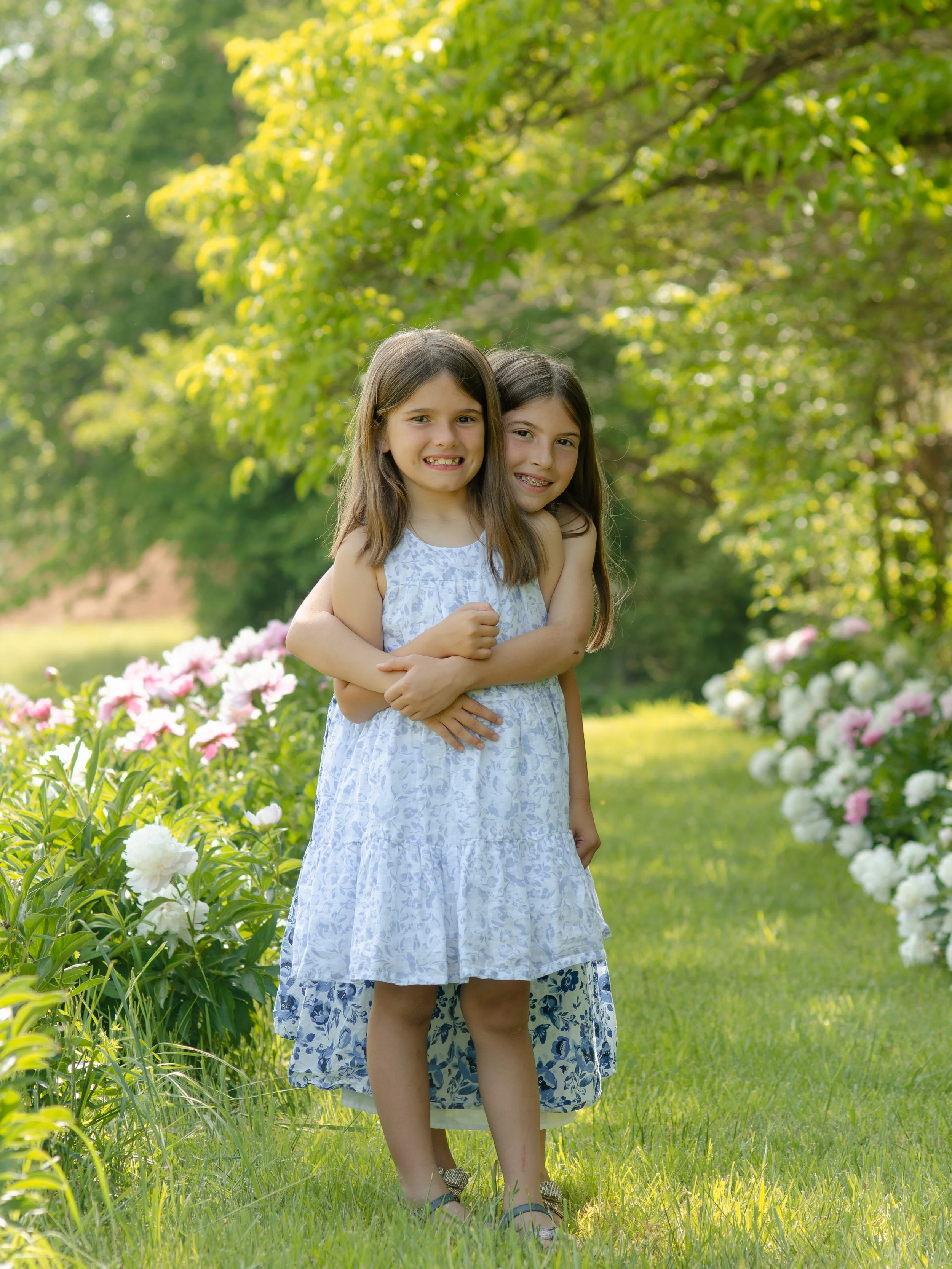 Playful sister photo session in Knoxville with happy interaction in flower field, smiling and embracing each other.