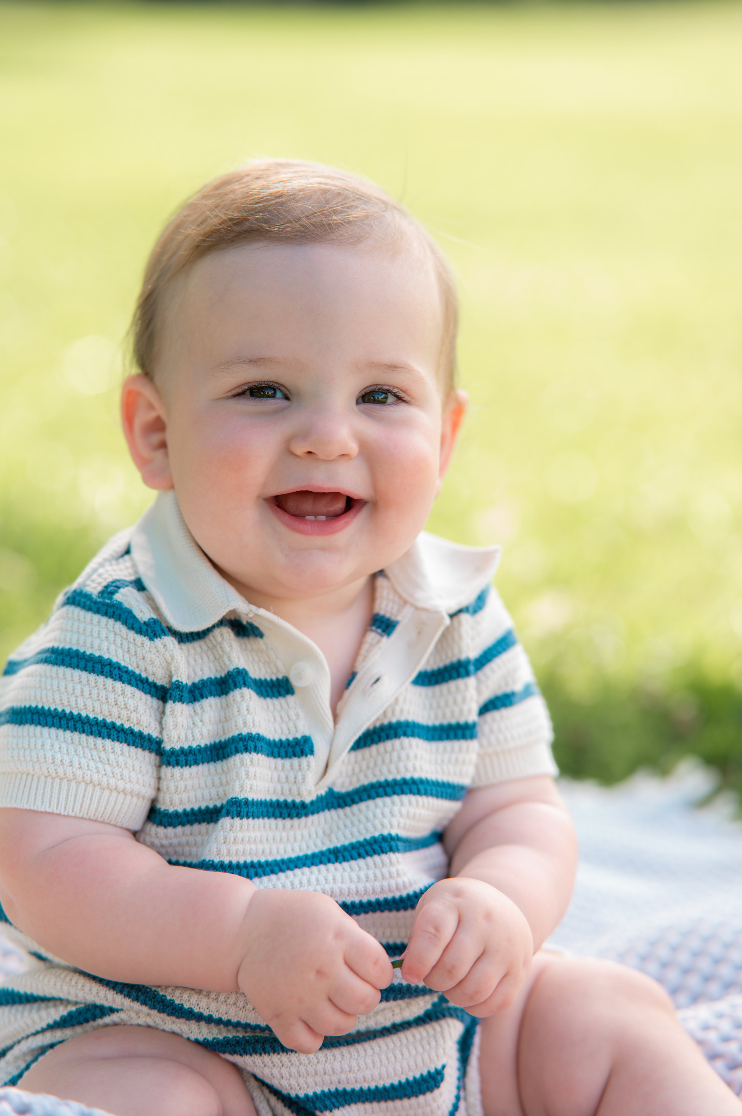 Close-up of a smiling baby boy with blue eyes and light brown hair, wearing a white and blue striped shirt, sitting outdoors on a sunny day.
