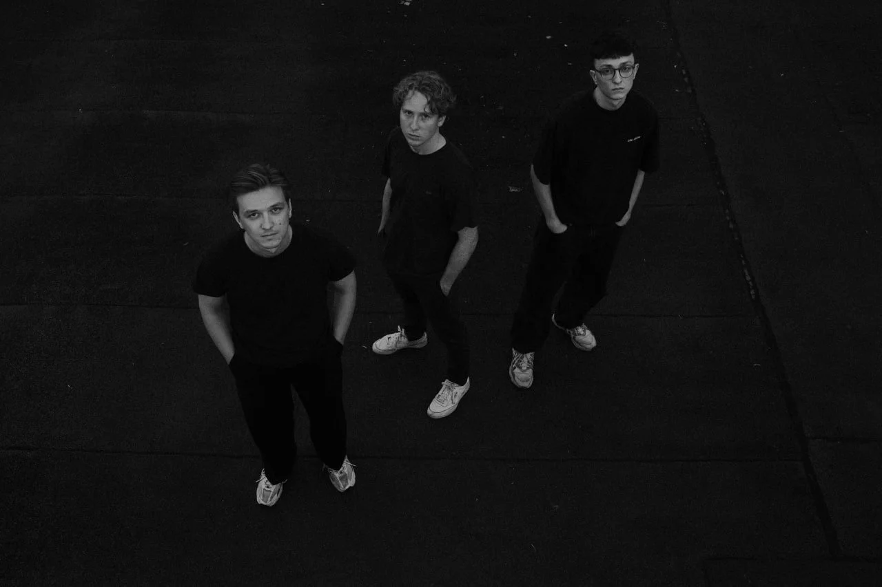 Three young men standing on a dark floor, looking up at the camera in black and white.