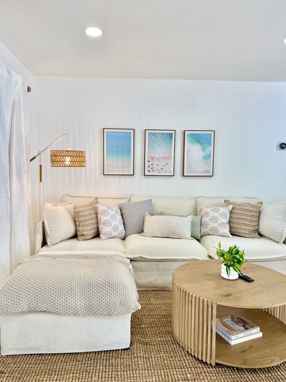 Living room with a white sofa, decorative pillows, a wooden coffee table with magazines and a potted plant, wall art with beach scenes, a tall floor lamp with a woven shade, and a jute rug.