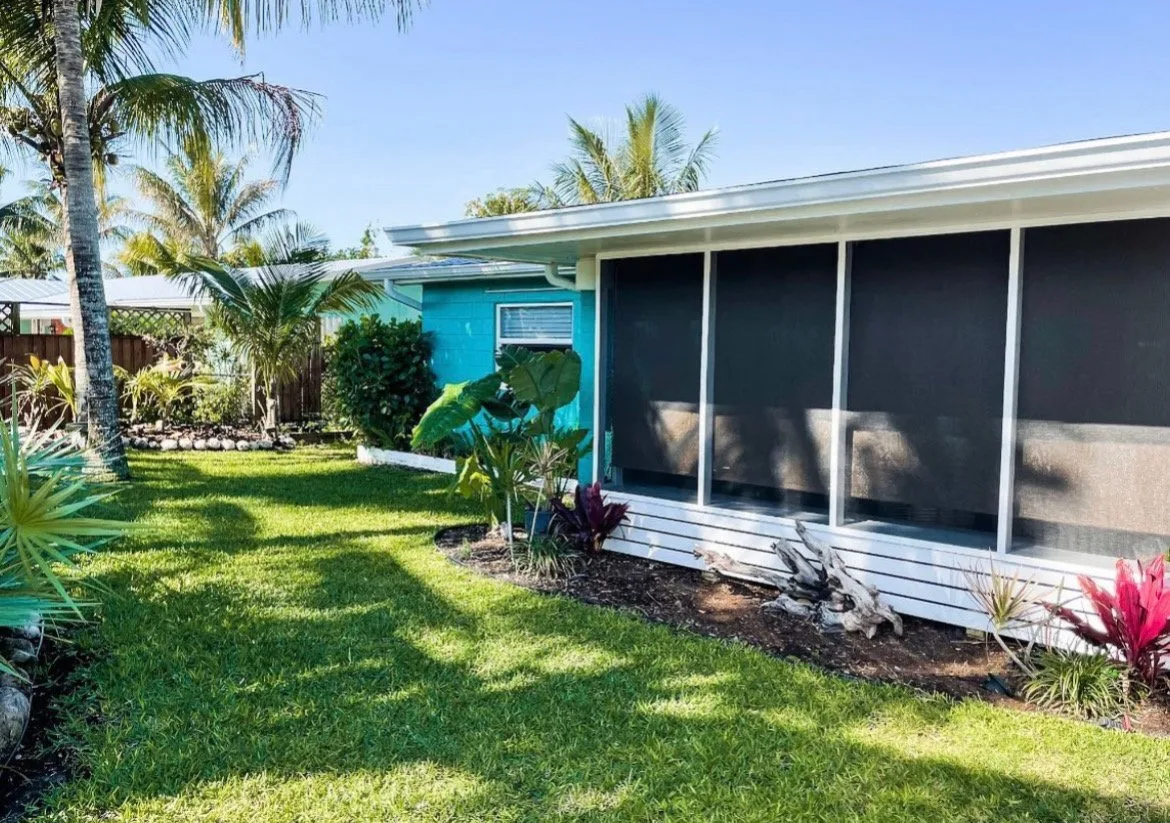 A backyard with green grass, tropical trees, and plants. There is a small house or shed painted in bright blue with black screened windows, and a white roof. The yard is sunny with clear blue sky.