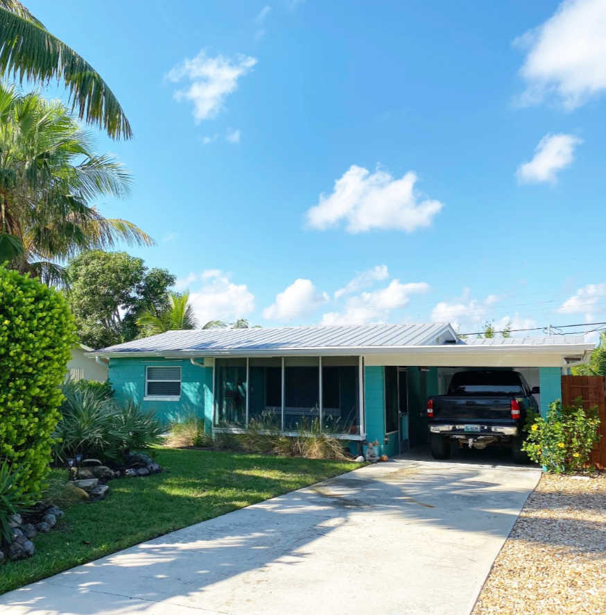 A turquoise house with a metal roof, a driveway with a black truck parked under a carport, and tropical landscaping including palm trees and bushes on a sunny day.